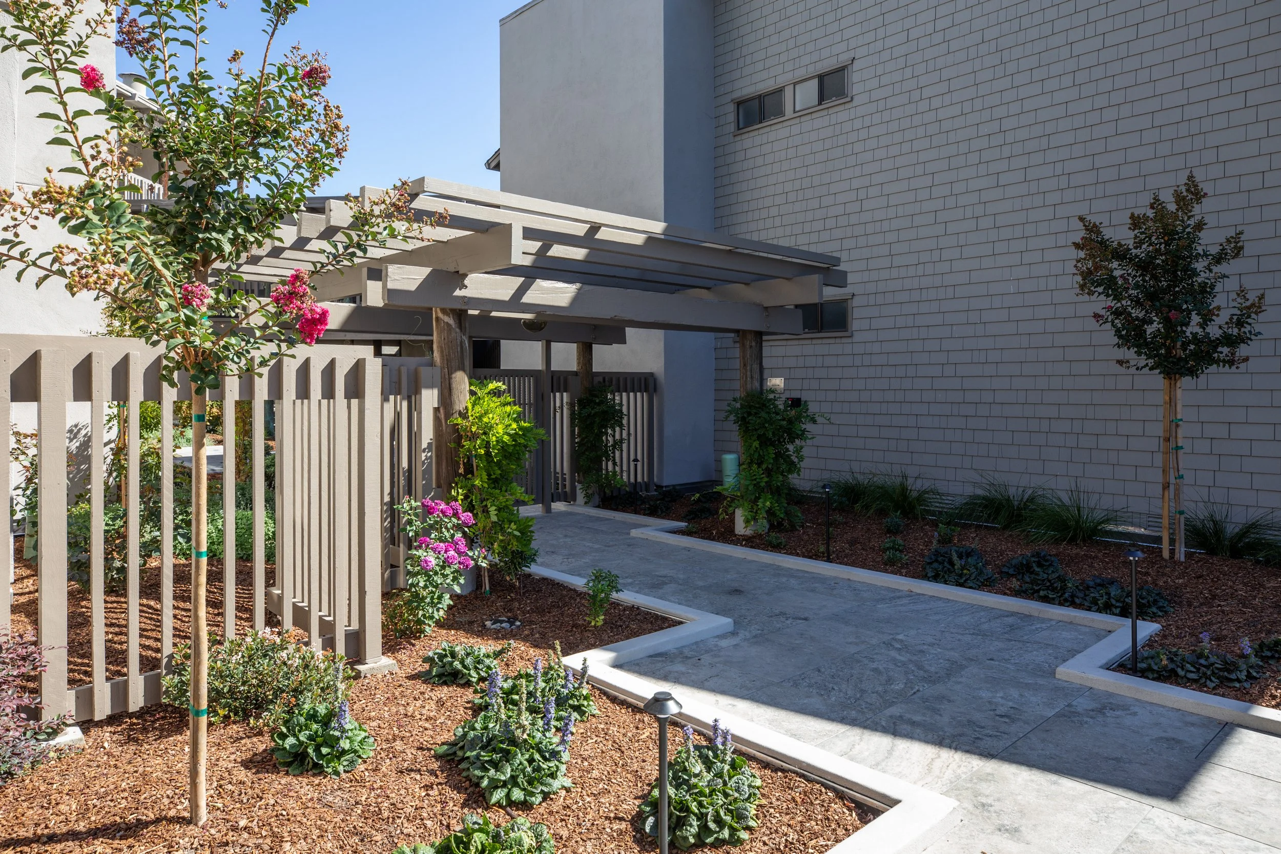 A modern building with a white brick exterior and small rectangular windows. In front, there is a garden with flowering plants, shrubs, and small trees, bordered by a beige picket fence and a concrete pathway.