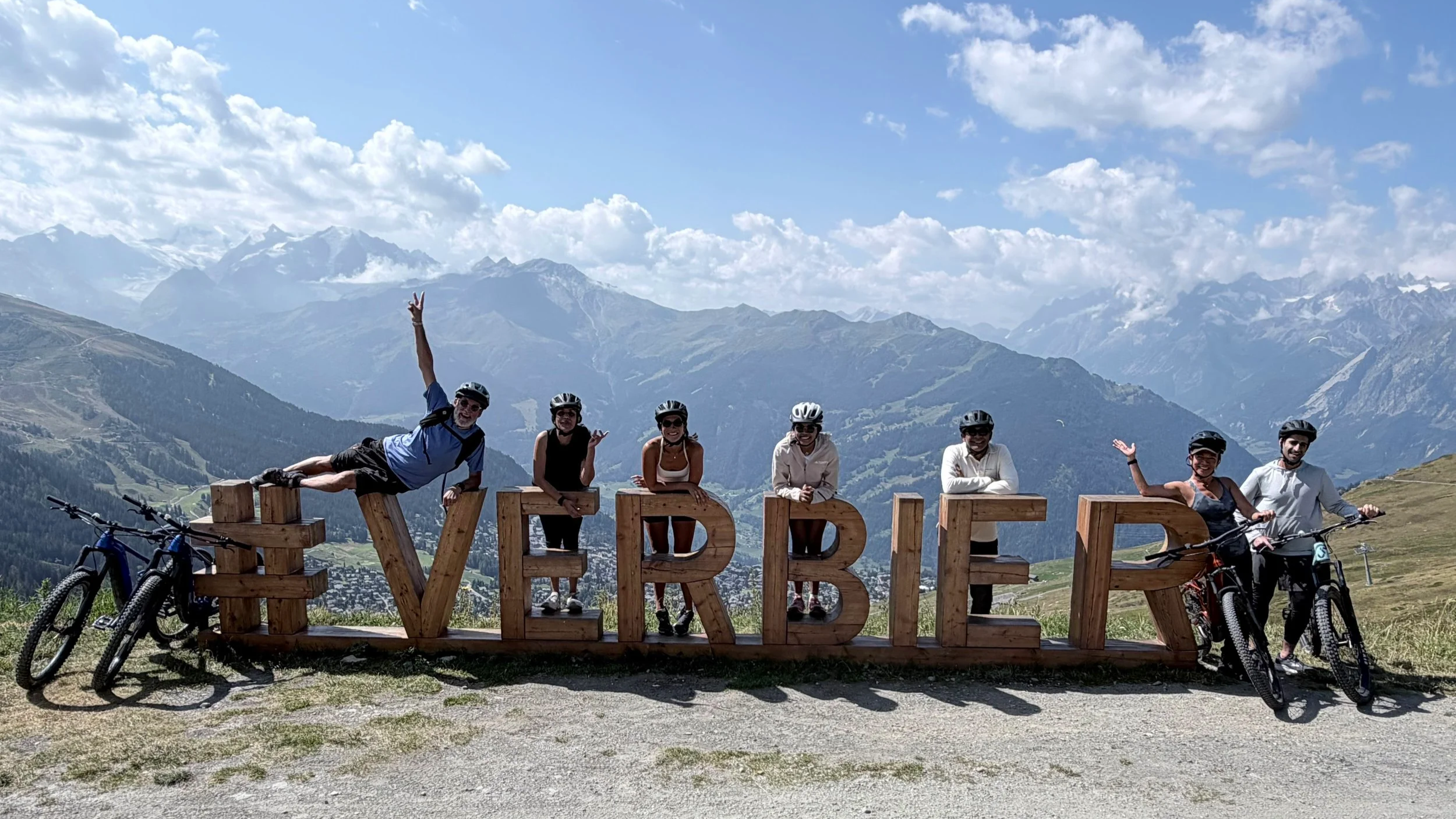 Group of six people, some wearing helmets, posing behind wooden letters spelling 'VERBIER' on a mountain landscape with snow-capped peaks and clouds in the sky, their bicycles are parked nearby.