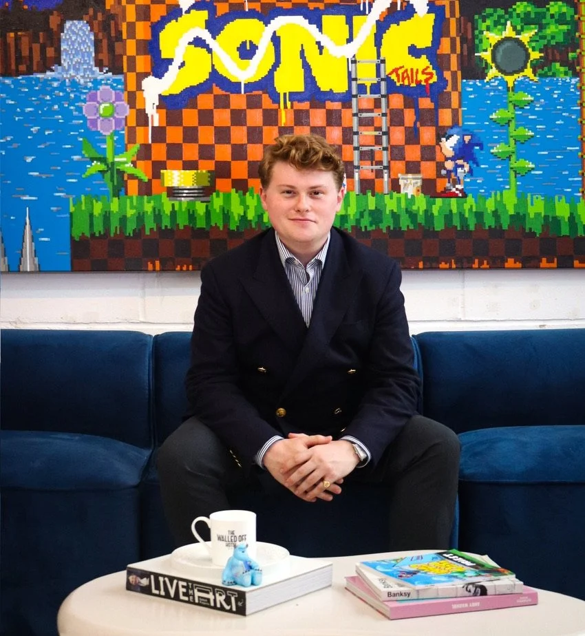 A young man sitting on a blue couch in front of a colorful pixel art mural that features the word 'SONIC' and scenes from the Sonic the Hedgehog video game, including green grass, blue water, and a checkered background.