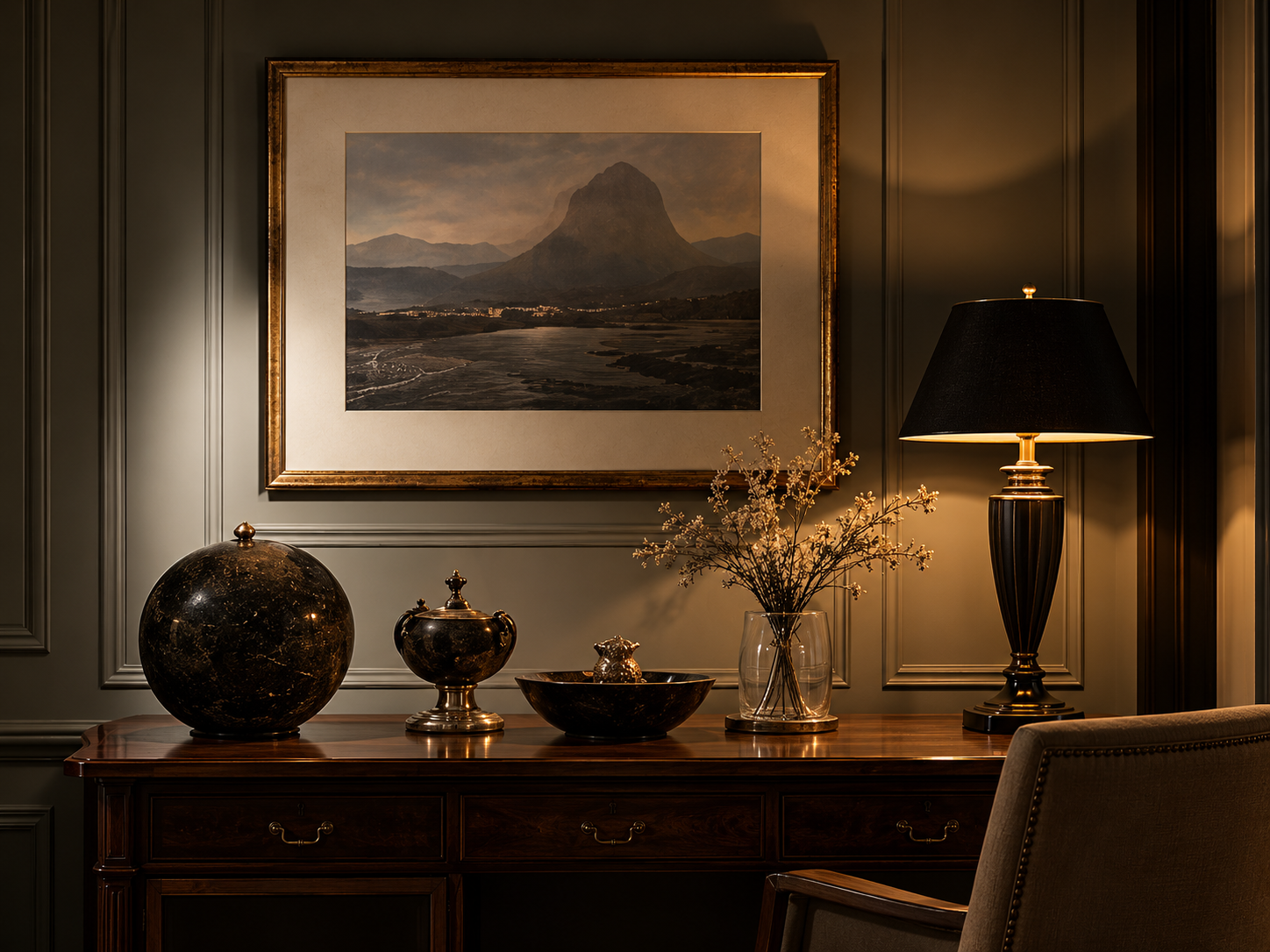 A dimly lit, elegant living room corner with a wooden sideboard, decorative vases, a glass vase with flowers, a framed picture of a mountain, and a black table lamp with a beige shade, all against a paneled wall.