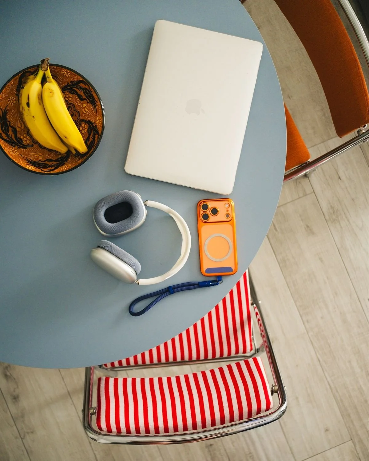 Mesa con una laptop blanca, un par de bananas en un plato decorativo, auriculares blancos y grises, y un teléfono móvil con funda naranja. En la esquina hay una silla con asiento de rayas rojas y blancas.