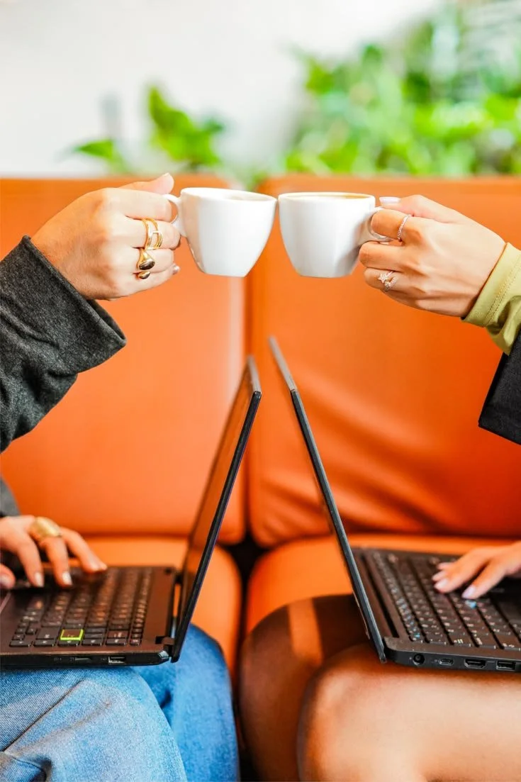 Dos personas intercambiando tazas de café o té mientras trabajan en laptops en un ambiente cómodo y luminoso.