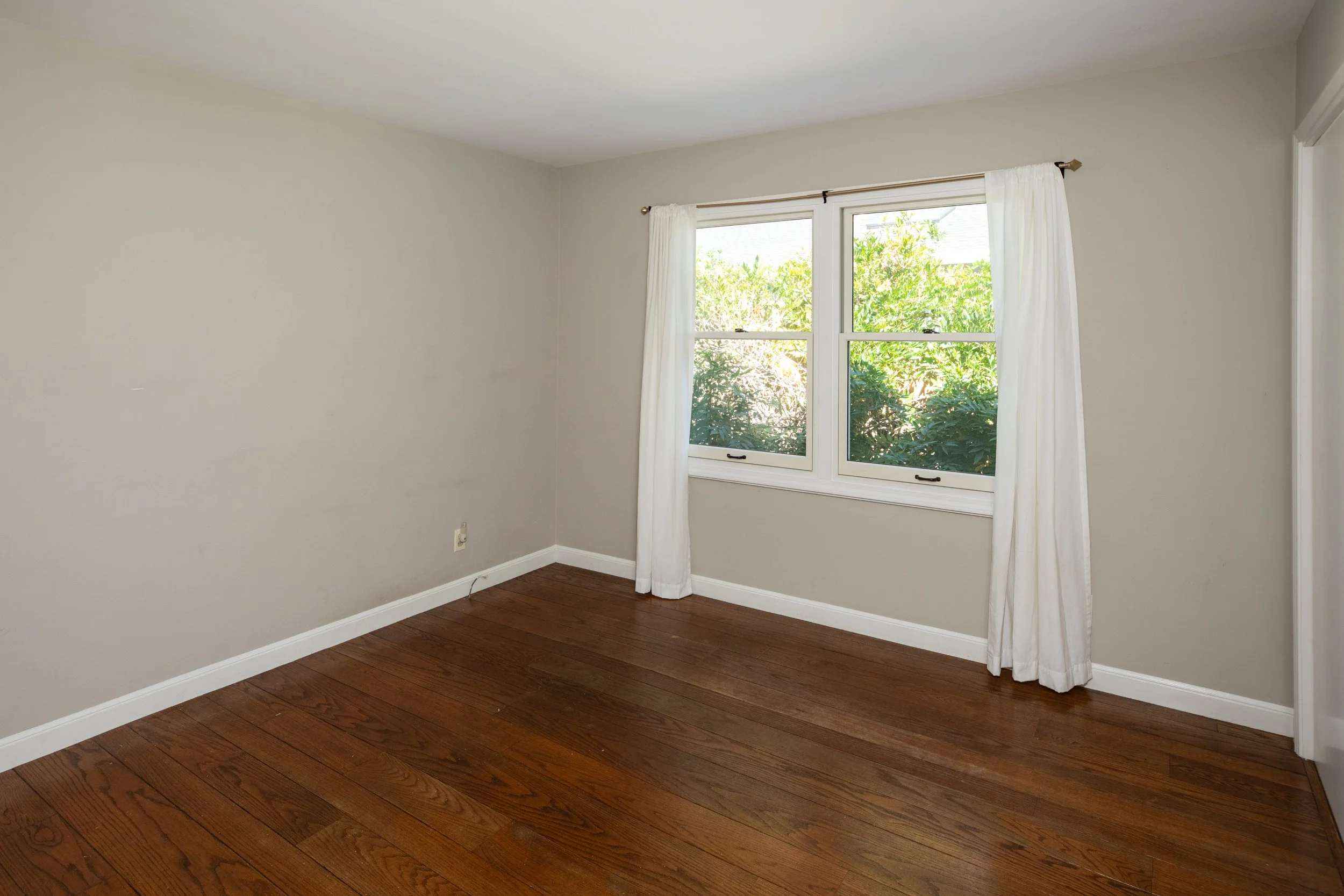Empty room with beige walls, hardwood flooring, a window with white curtains, and outside greenery visible through the window.
