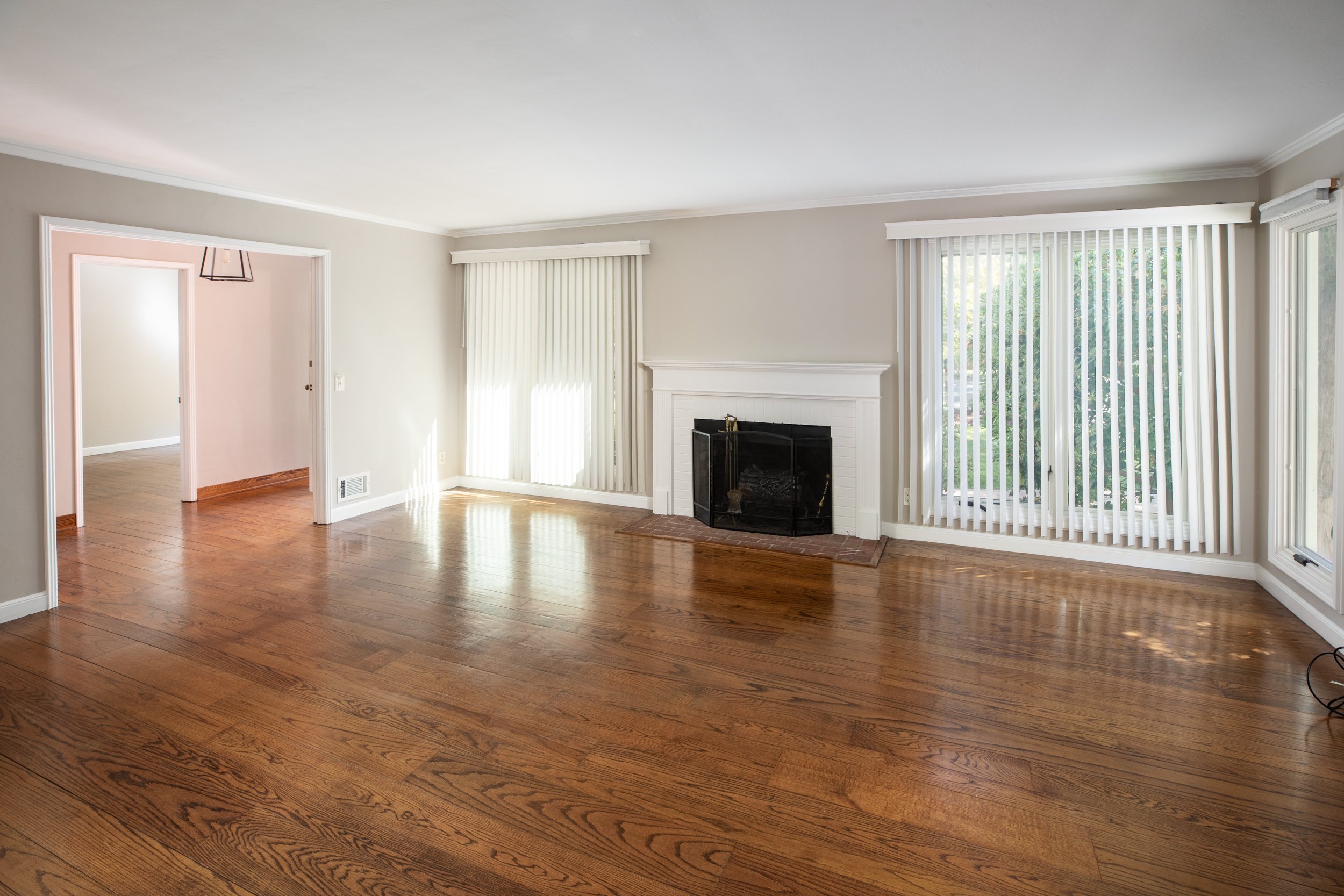 Living room with hardwood floors, large windows with vertical blinds, a white fireplace, and doorway leading to another room.