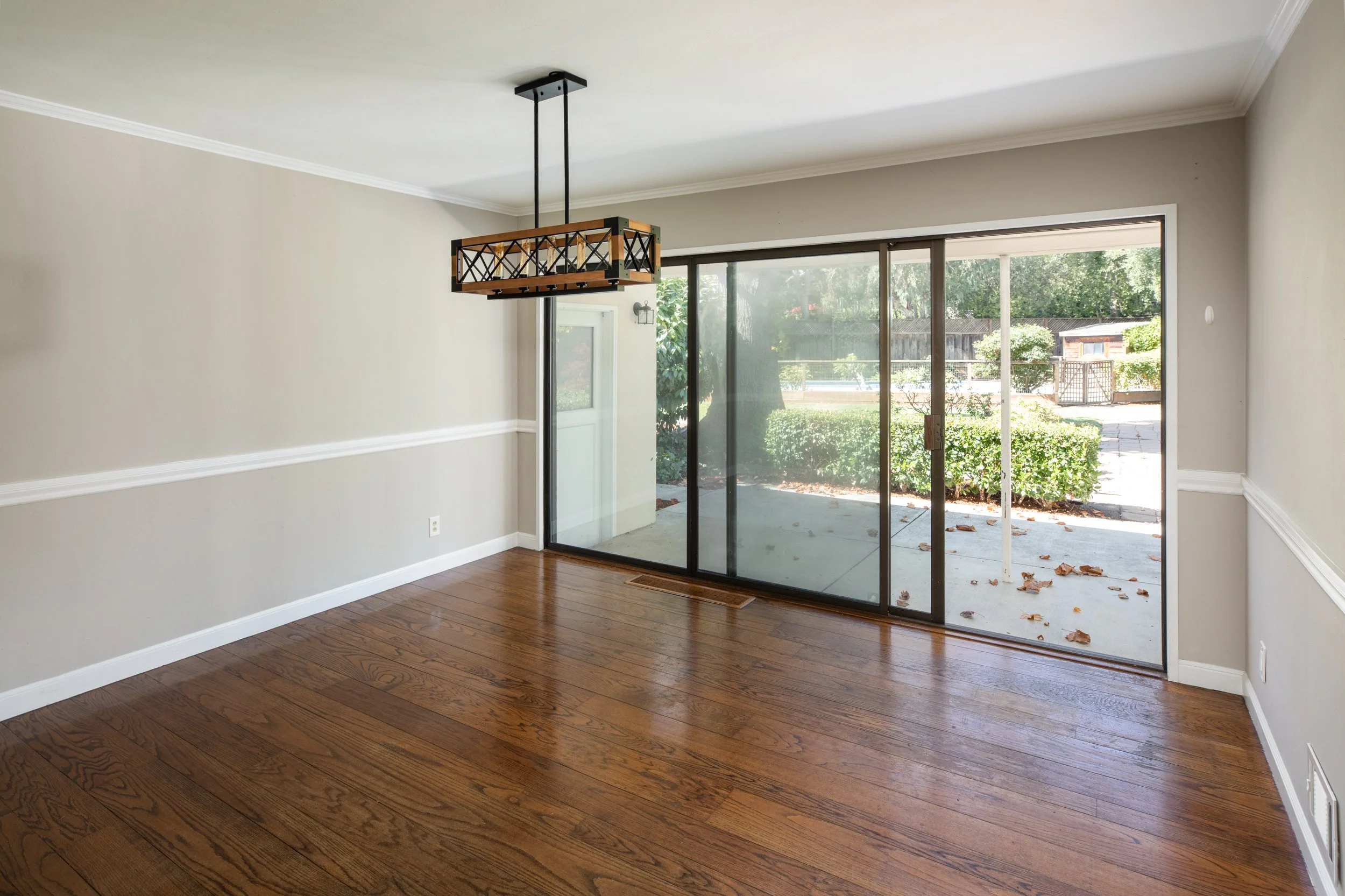 Interior view of a room with hardwood floors, a beige wall with white trim, a black and wood chandelier, and a large sliding glass door leading outside to a patio and yard with bushes and trees.
