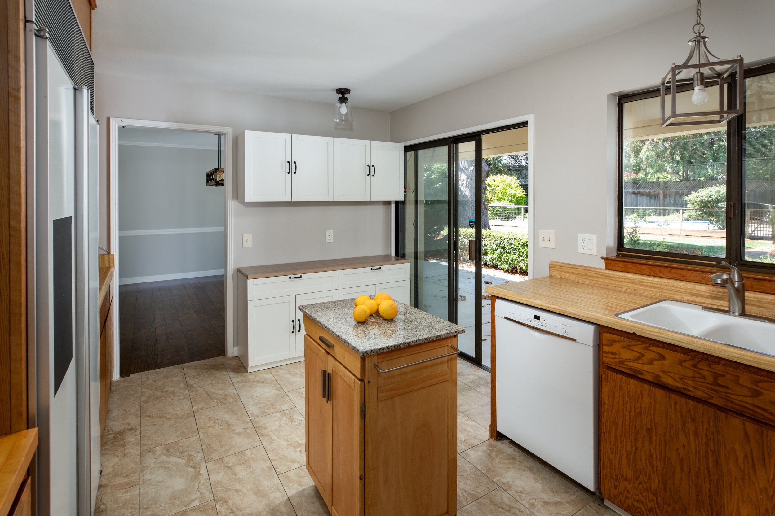 A kitchen with beige tiled floor, white walls, white cabinets, a wooden counter, and a sliding glass door leading outside. There is a small wooden island with lemons on top, and a window above the sink showing a yard with trees.