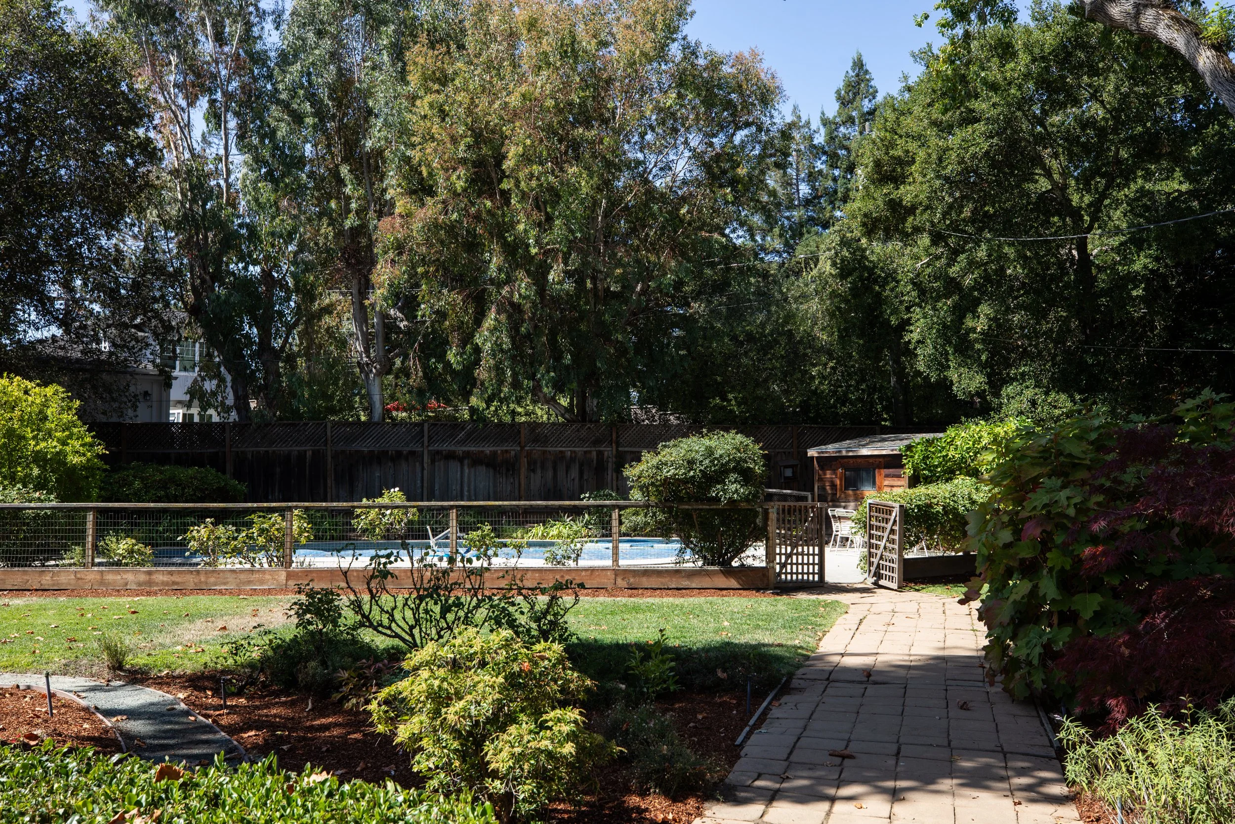 A backyard garden with a wooden fence, trees, shrubs, and a small patio area with chairs and a table. A swimming pool is visible behind the fence.