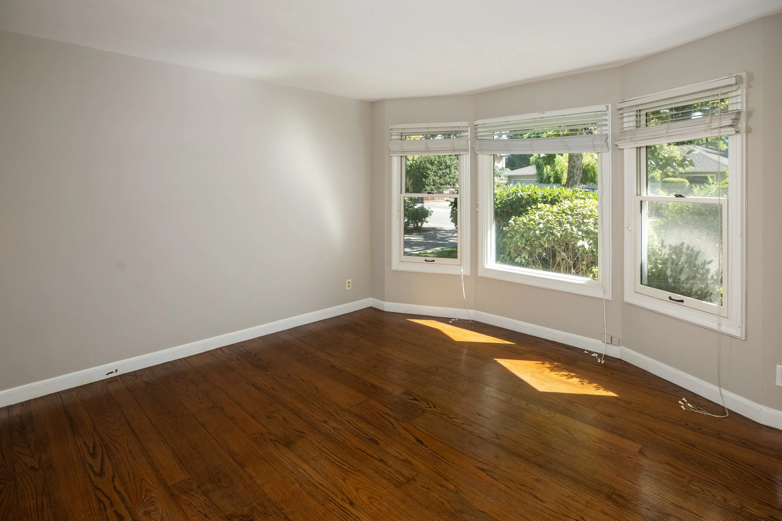 Empty room with hardwood floors, white walls, and three large windows with blinds showing trees and neighboring houses outside.