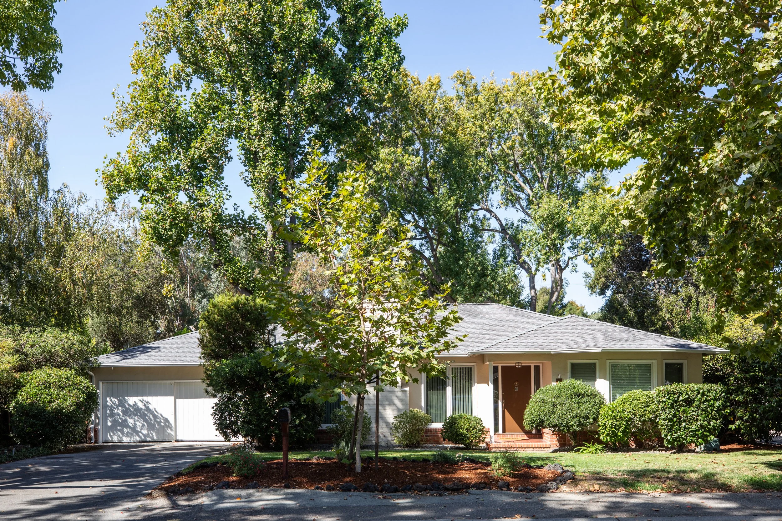 Front view of a suburban house with a grey roof, surrounded by green trees and bushes, with a clear blue sky.