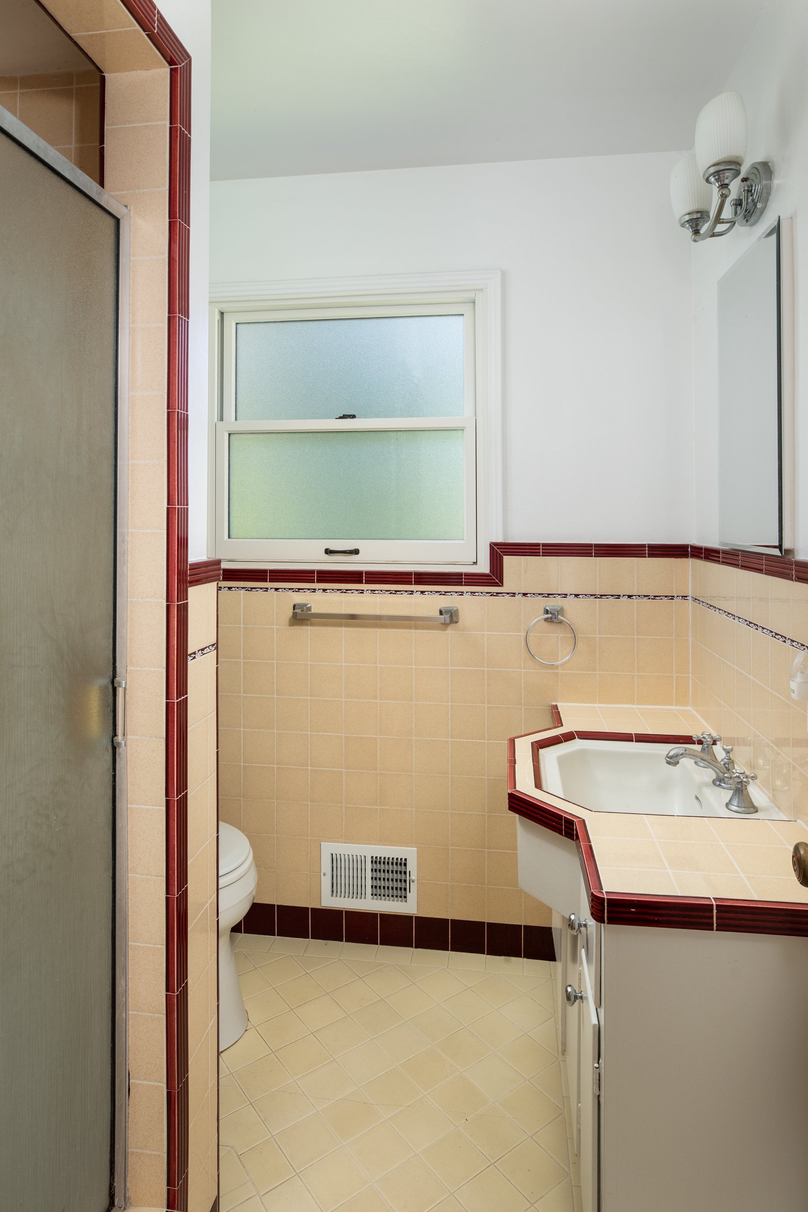 Bathroom with beige tiles, red trim accents, a frosted window, a white toilet, a small corner sink with red tile border, and a wall-mounted mirror and light fixture.