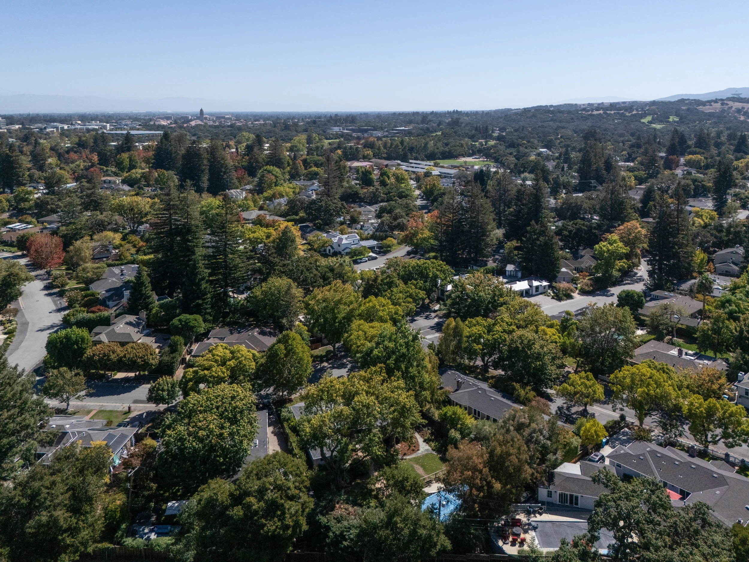 An aerial view of a residential neighborhood with numerous houses, trees, and streets, overlooking a distant city and hills under a clear blue sky.