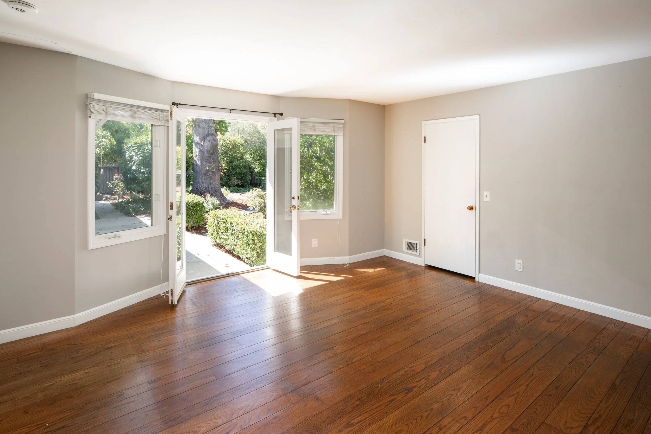 Empty room with hardwood flooring, white walls, and large windows with blinds, open to a garden with trees and bushes.
