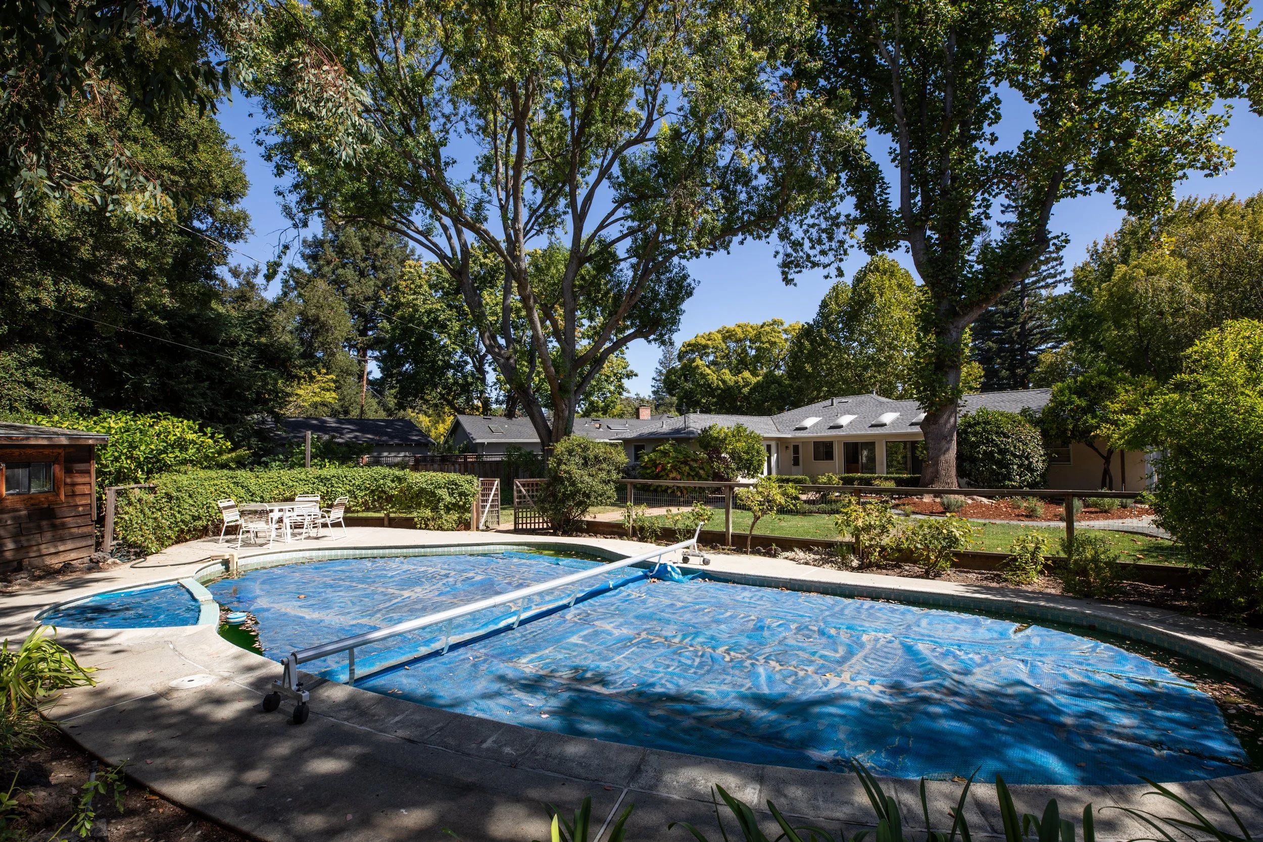 Backyard with a swimming pool covered by a blue tarp, surrounded by patio furniture, trees, and houses in the background under a sunny sky.