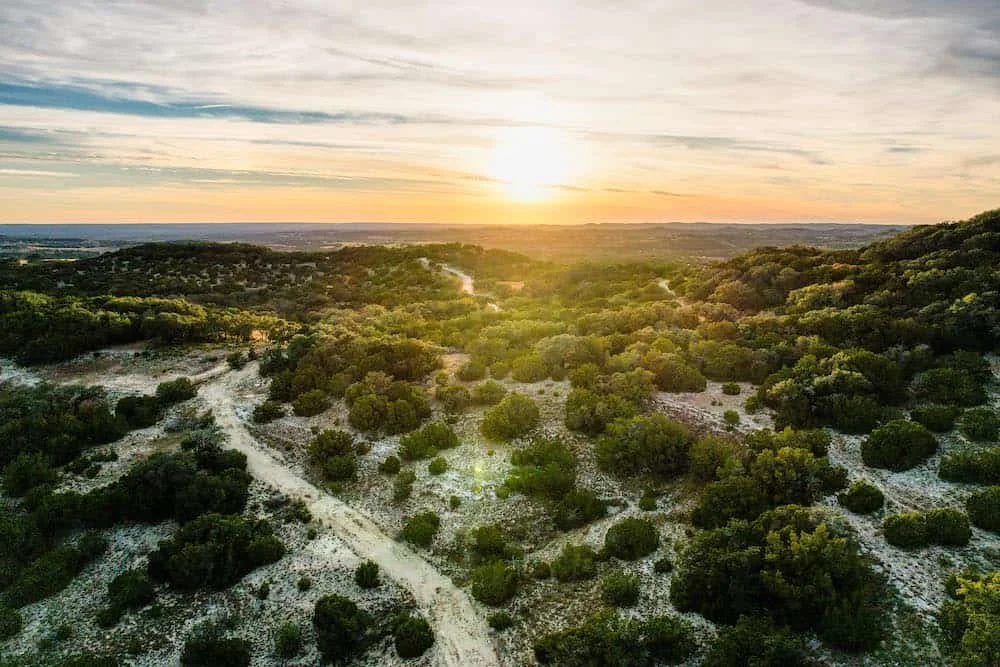 Aerial view of a landscape with dirt paths, green bushes, trees, and a sunset horizon.