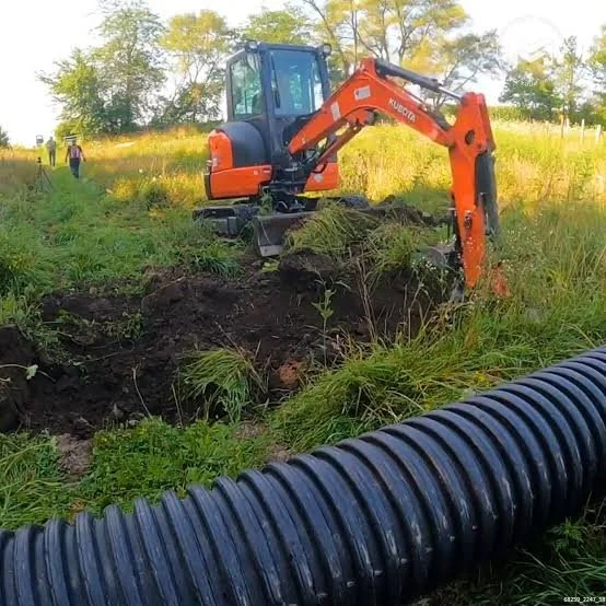 Small orange excavator digging a trench in a grassy field with trees in the background, and large black drainage pipe nearby.