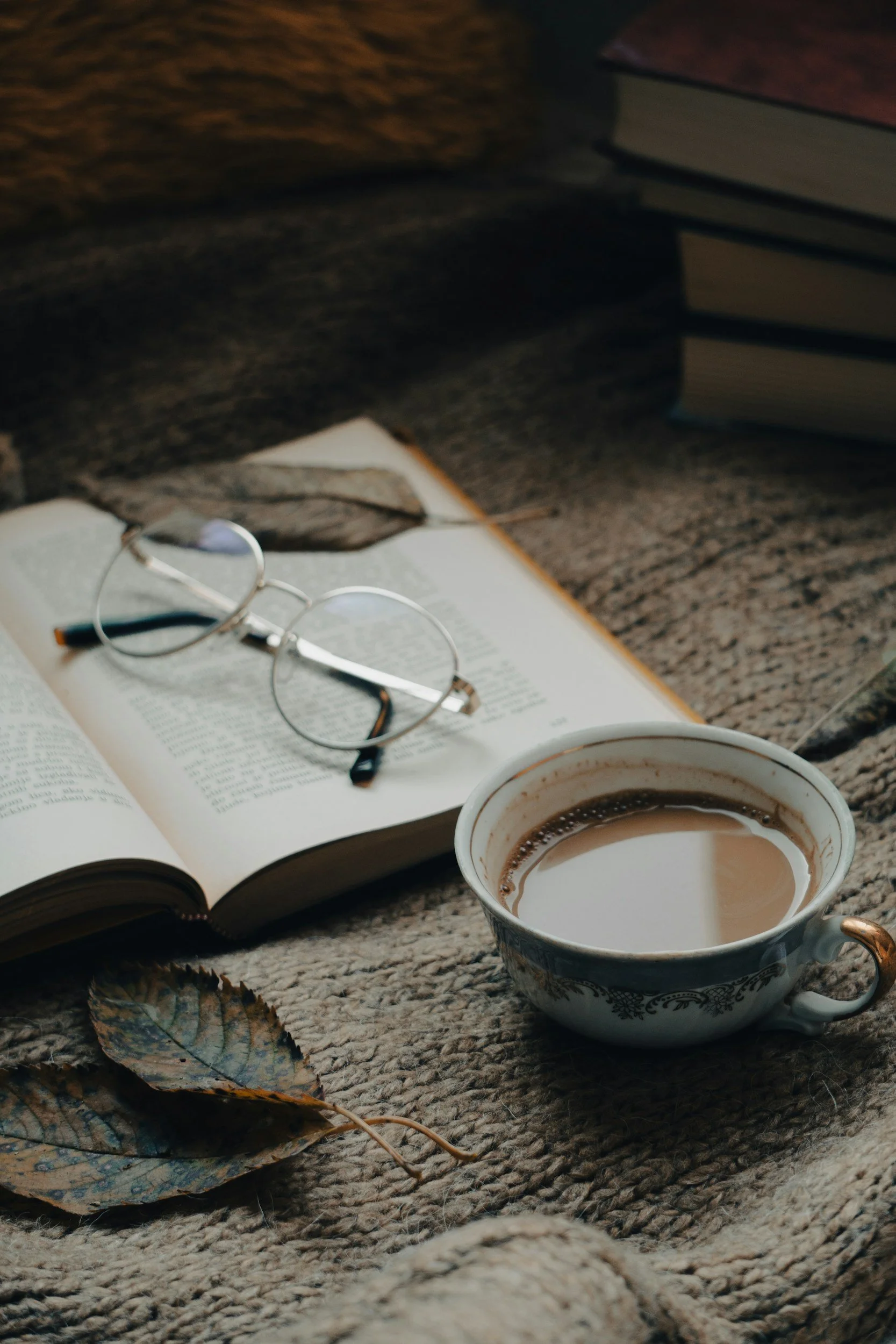 A cup of coffee placed on a textured fabric surface, with an open book and a pair of eyeglasses resting on the pages. In the background, there are stacked books and some fallen autumn leaves.