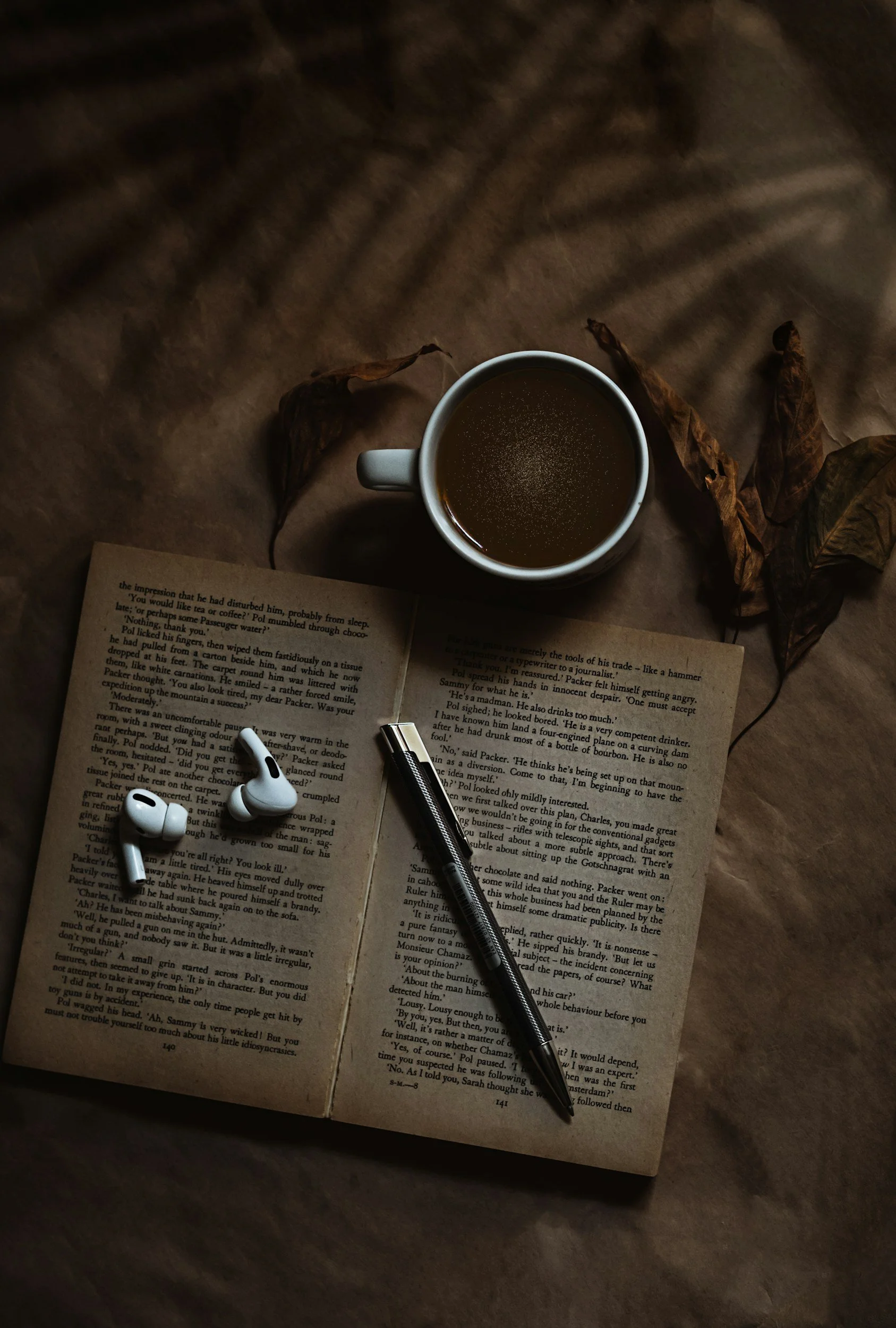 A top view of an open book, a black pen, a white mug filled with dark coffee, a pair of white wireless earbuds, and dried autumn leaves on a wooden surface.