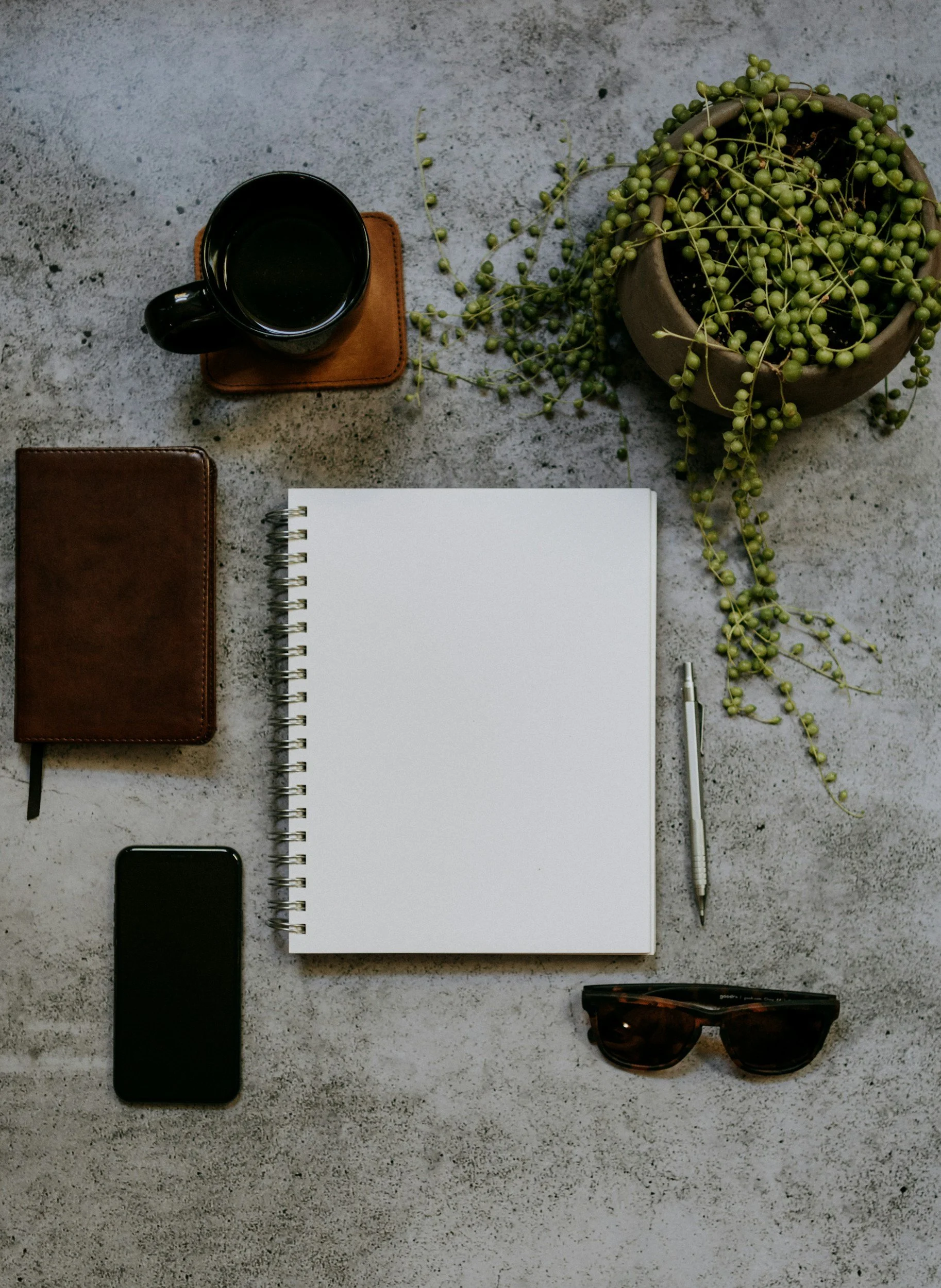 A top-down view of a workspace with a coffee mug on a leather coaster, a closed leather notebook, a spiral notebook with blank pages, a smartphone, a pen, a pair of sunglasses, and a potted plant with green berries on a concrete surface.