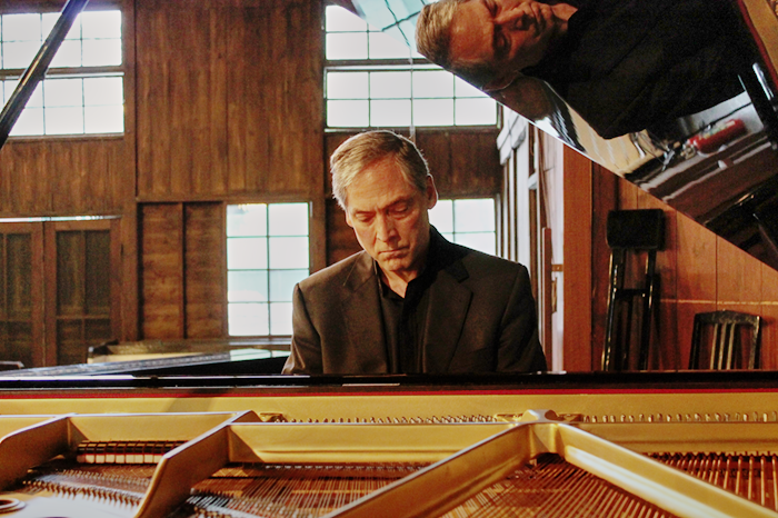 Older man in a black suit playing a grand piano inside a wood-paneled room