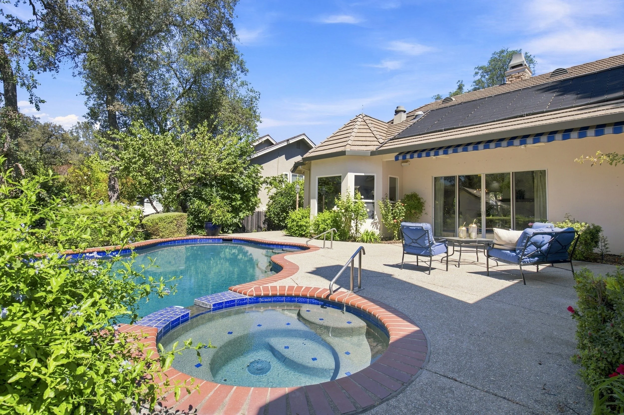 Residential backyard with a small swimming pool, patio furniture, lush greenery, and a house with solar panels on the roof.