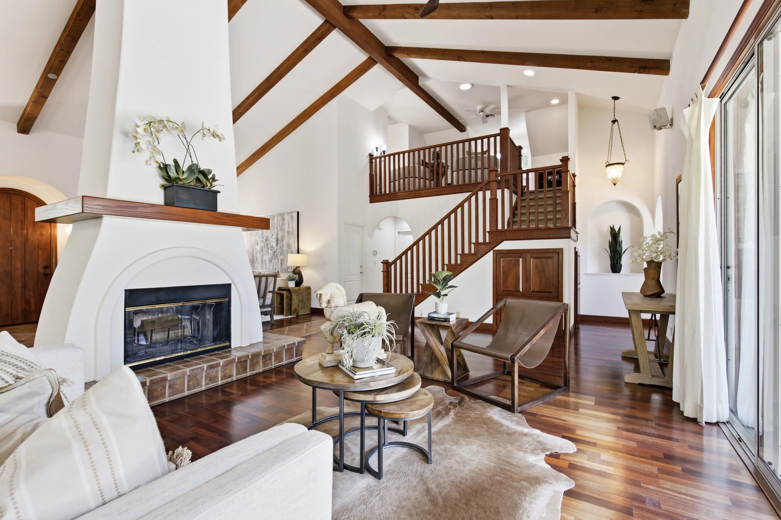 Interior view of a spacious living room with a high, vaulted ceiling with wooden beams, a white fireplace with a mantel decorated with plants, wooden staircase and railing, large windows with curtains, wood flooring, and contemporary furniture includ