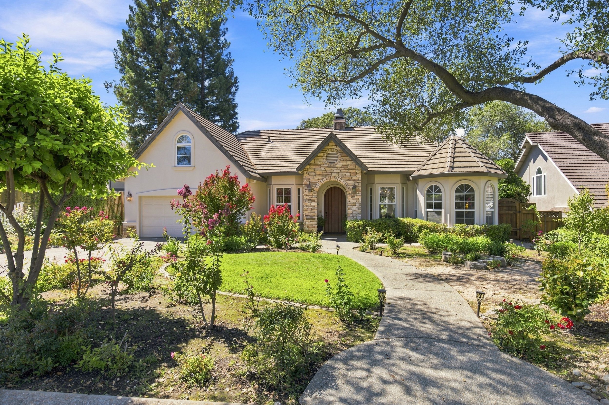 A suburban house with a curved front lawn and garden, surrounded by trees and plants, under a partly cloudy blue sky.