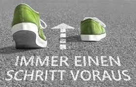 Zwei grüne Turnschuhe auf einer Straße, mit Pfeil und theatralischer Beschriftung "Immer einen Schritt voraus" in deutscher Sprache