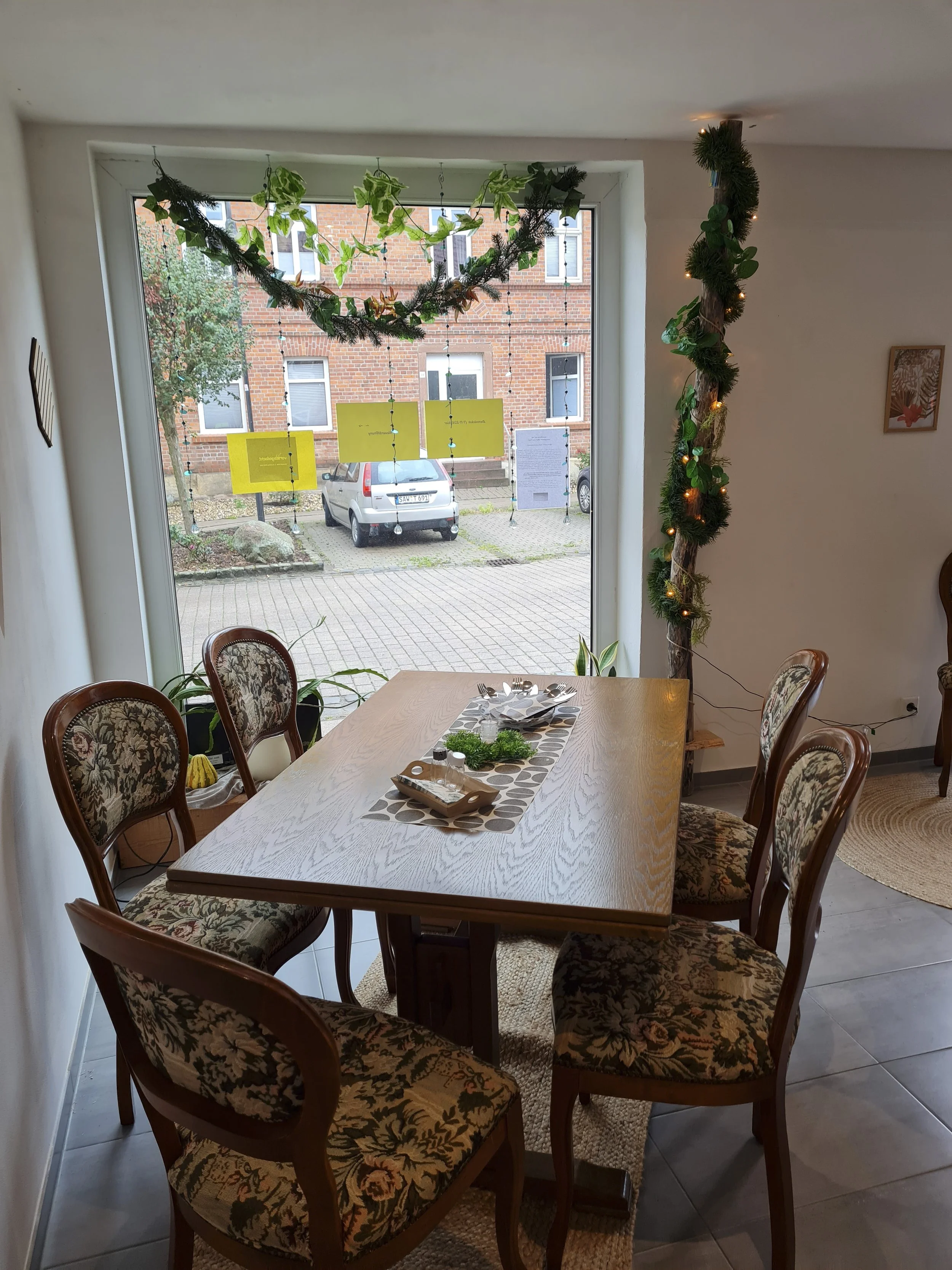 Innenaufnahme eines Esszimmers mit einem Holztisch, sechs gepolsterten Stühlen in Blumenmuster, ein Fenster mit grünen Pflanzen und Dekorationen, eine Wand mit eingerahmten Bildern, Blick nach draußen auf einen Parkplatz mit Autos und einem Backsteinhaus.