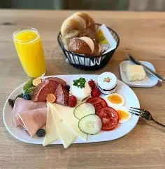 Frühstücksteller mit Wurst, Käse, Tomaten, Gurken, Rucola, Butter, Salz, Pfeffer, dazu ein Glas Orangensaft und eine Schüssel mit Brotkugeln auf einem Holztisch.