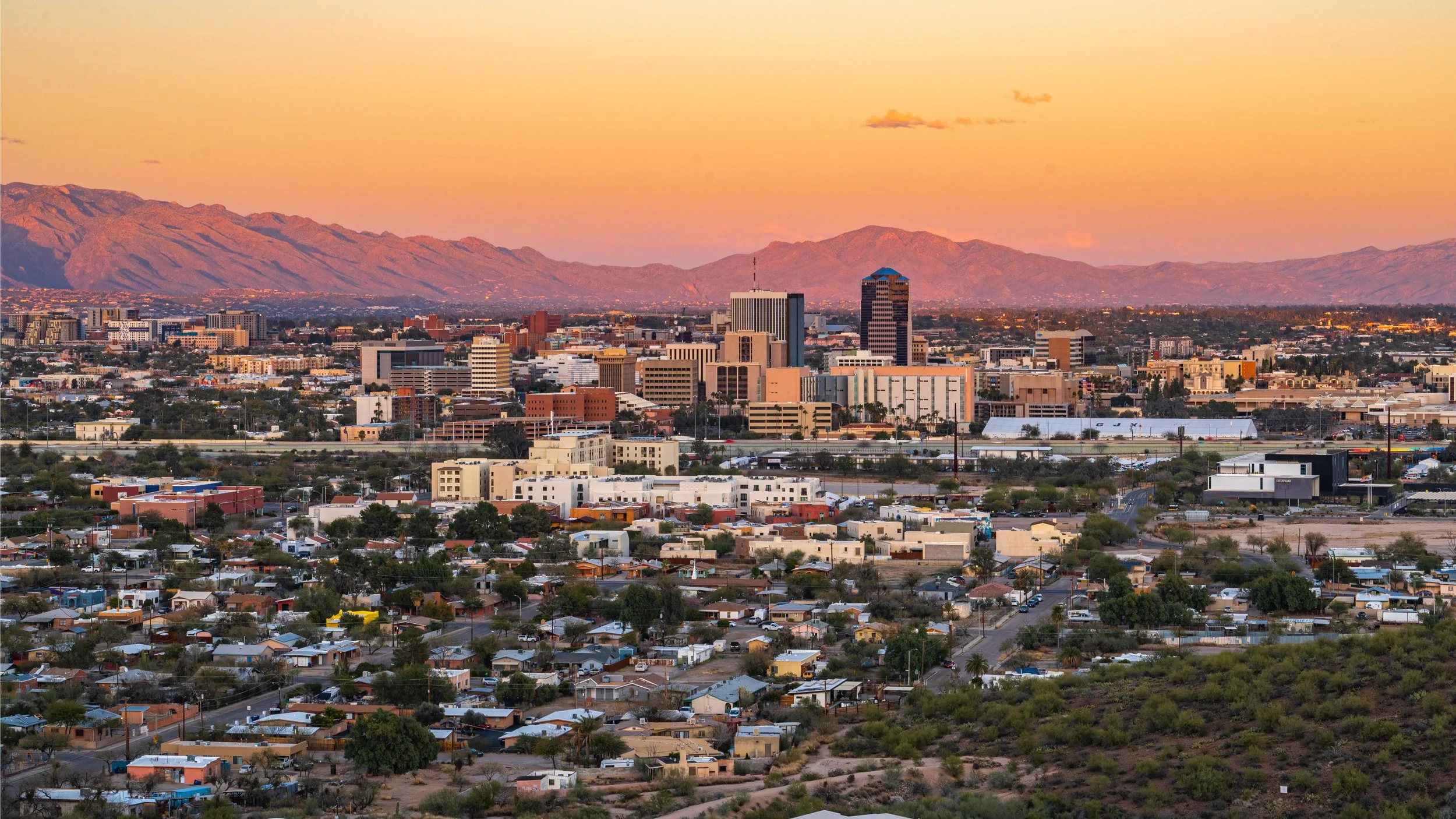 Downtown Tucson skyline and surrounding neighborhoods beneath an orange desert sunset and mountain backdrop.