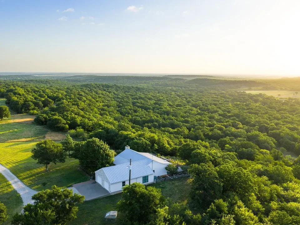 A white house surrounded by green trees, with a driveway and open fields nearby, under a clear sky at sunset.