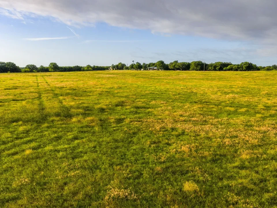 Open grassy field with tire tracks, distant trees, and a few buildings under a blue sky with some clouds.