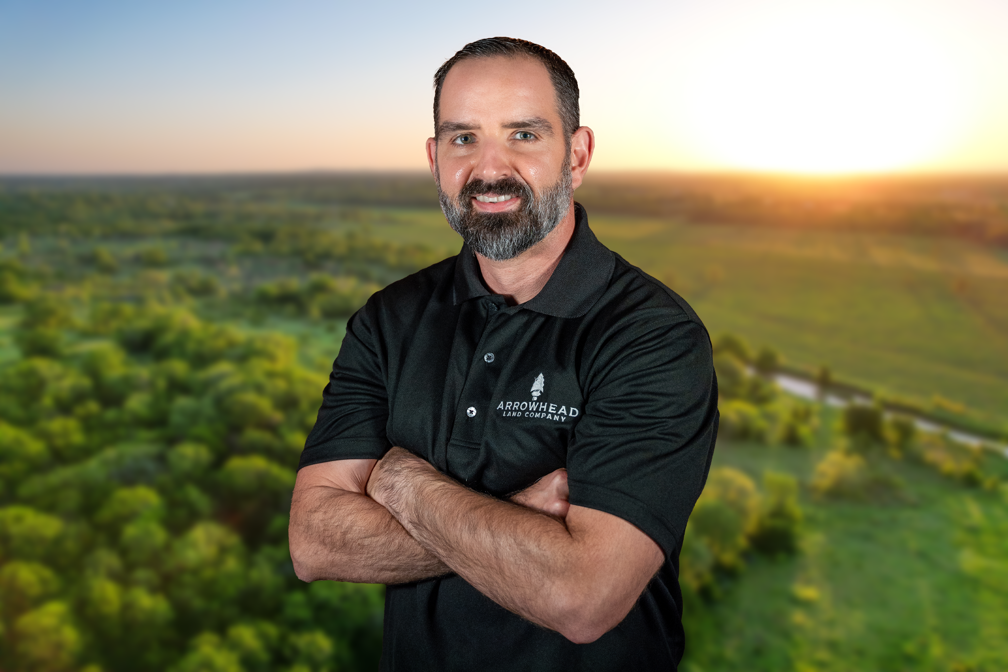 A man with a beard and mustache, wearing a black polo shirt with the logo 'Arrowhead Land Company', standing with his arms crossed in front of a blurred landscape background at sunset.