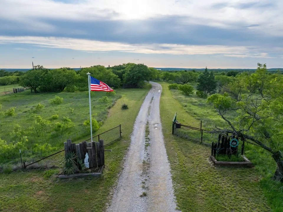 A gravel driveway leading through a rural property with a flagpole flying the American flag. There are small gardens on each side of the entrance, with a tree on the right side and a sunset or sunrise sky overhead.