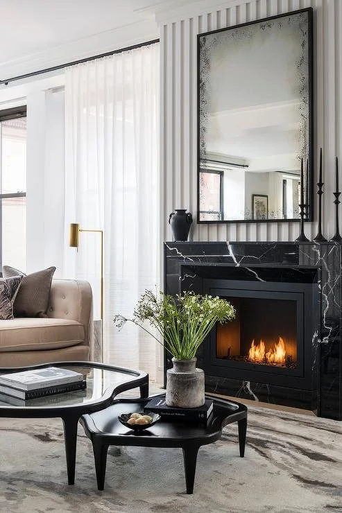 Modern living room with black marble fireplace, large mirror, beige sofa, glass coffee table, white curtains, and houseplants.