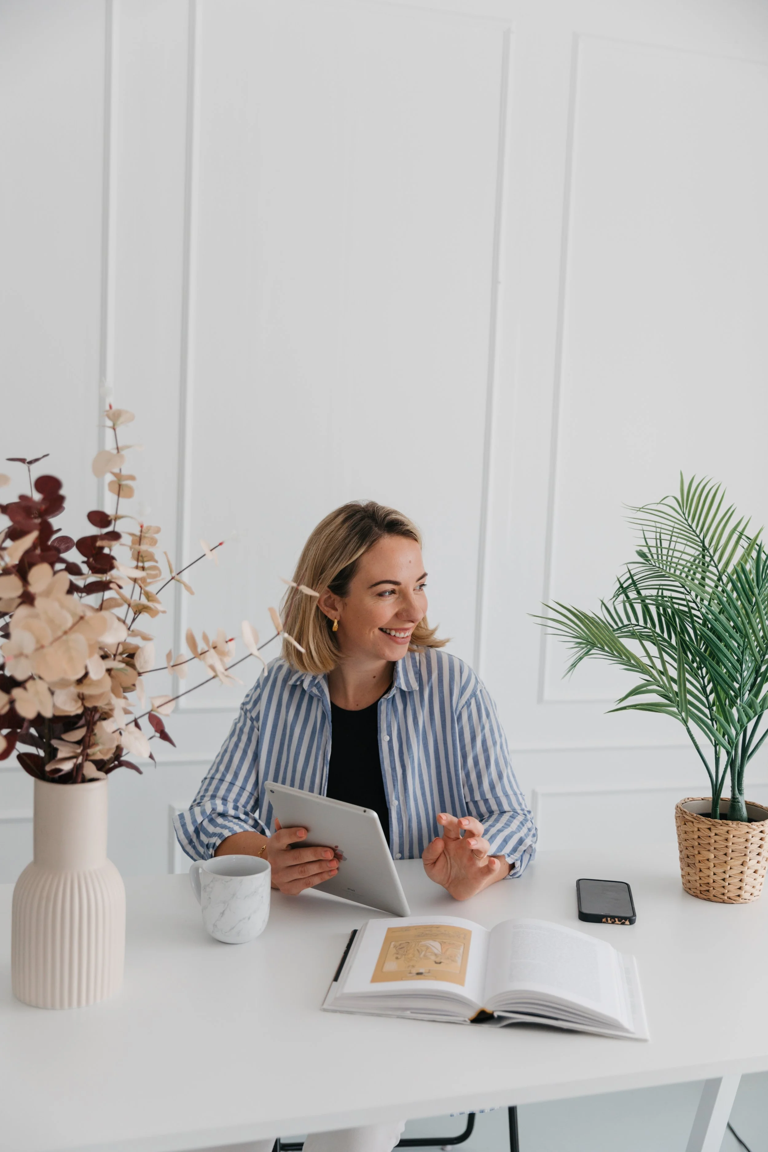 A woman sitting at a white table in a bright room, smiling and holding a tablet. The table has a large open book, a smartphone, a ceramic mug, and a white vase with dried flowers and green foliage.