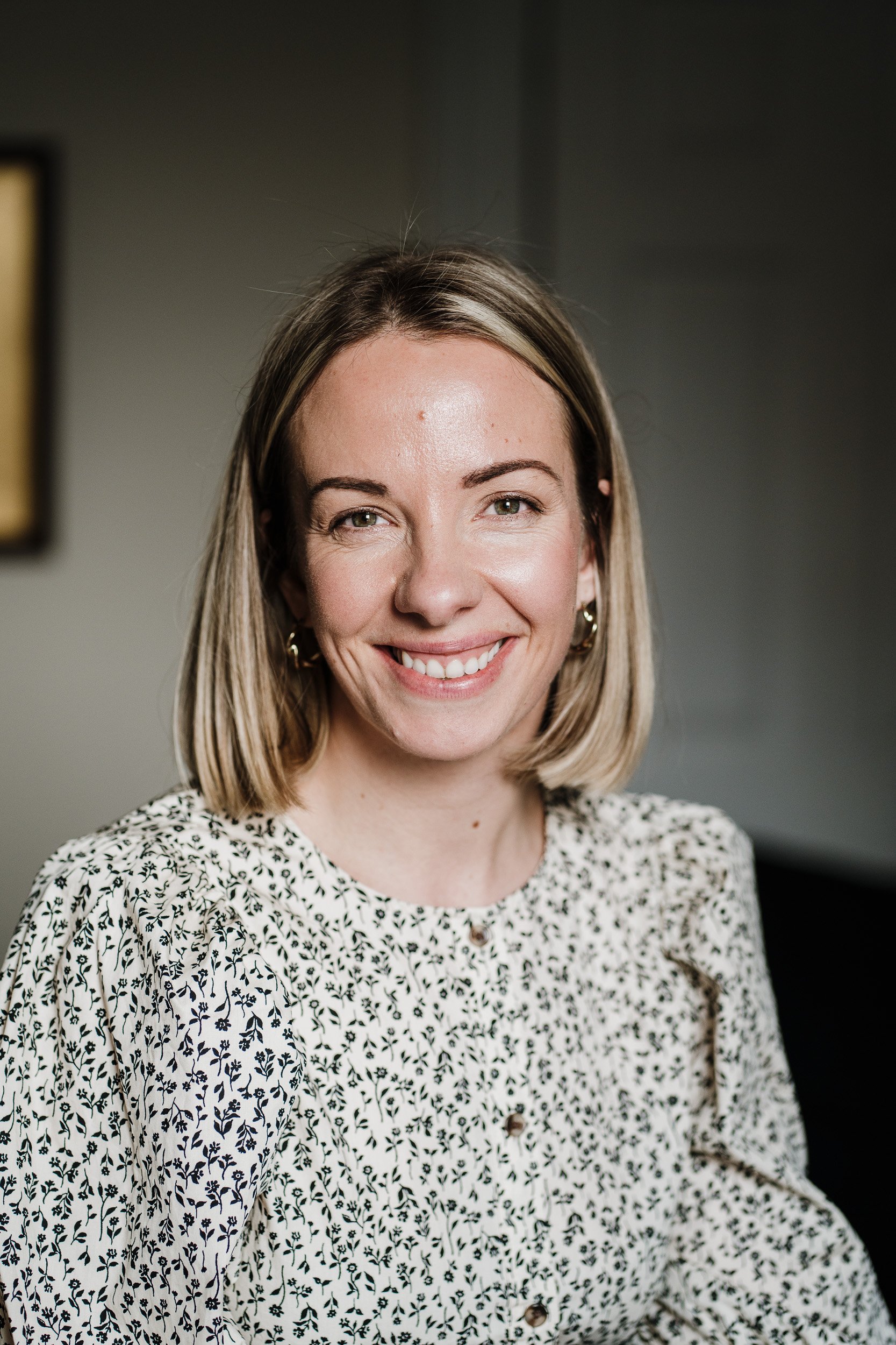 A woman with shoulder-length blonde hair smiling at the camera, wearing a white blouse with black leaf patterns, earrings, and sitting in an indoor setting.