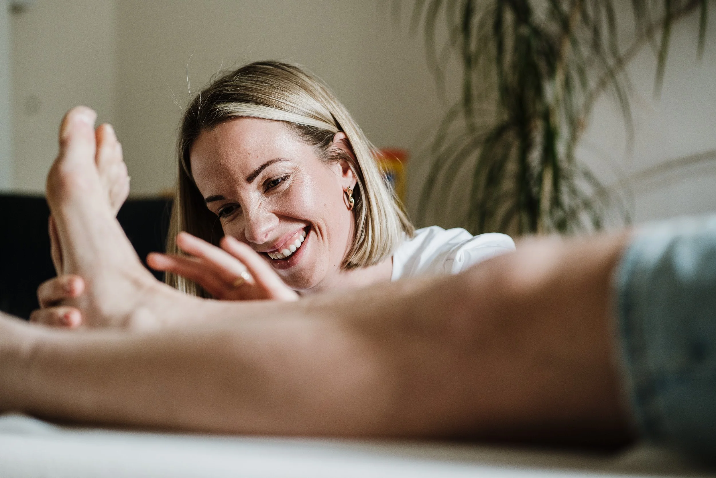 A woman with blonde hair smiling and engaging in a playful moment with another person's hand in a bright, indoor setting, with a large plant in the background.
