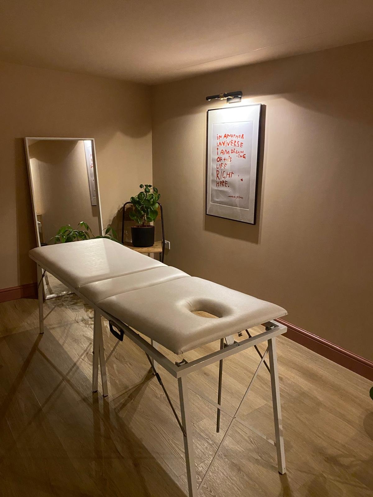 A massage room with a massage table covered with a white sheet, a large mirror, a potted plant on a small chair, a framed quote on the wall, and warm lighting.