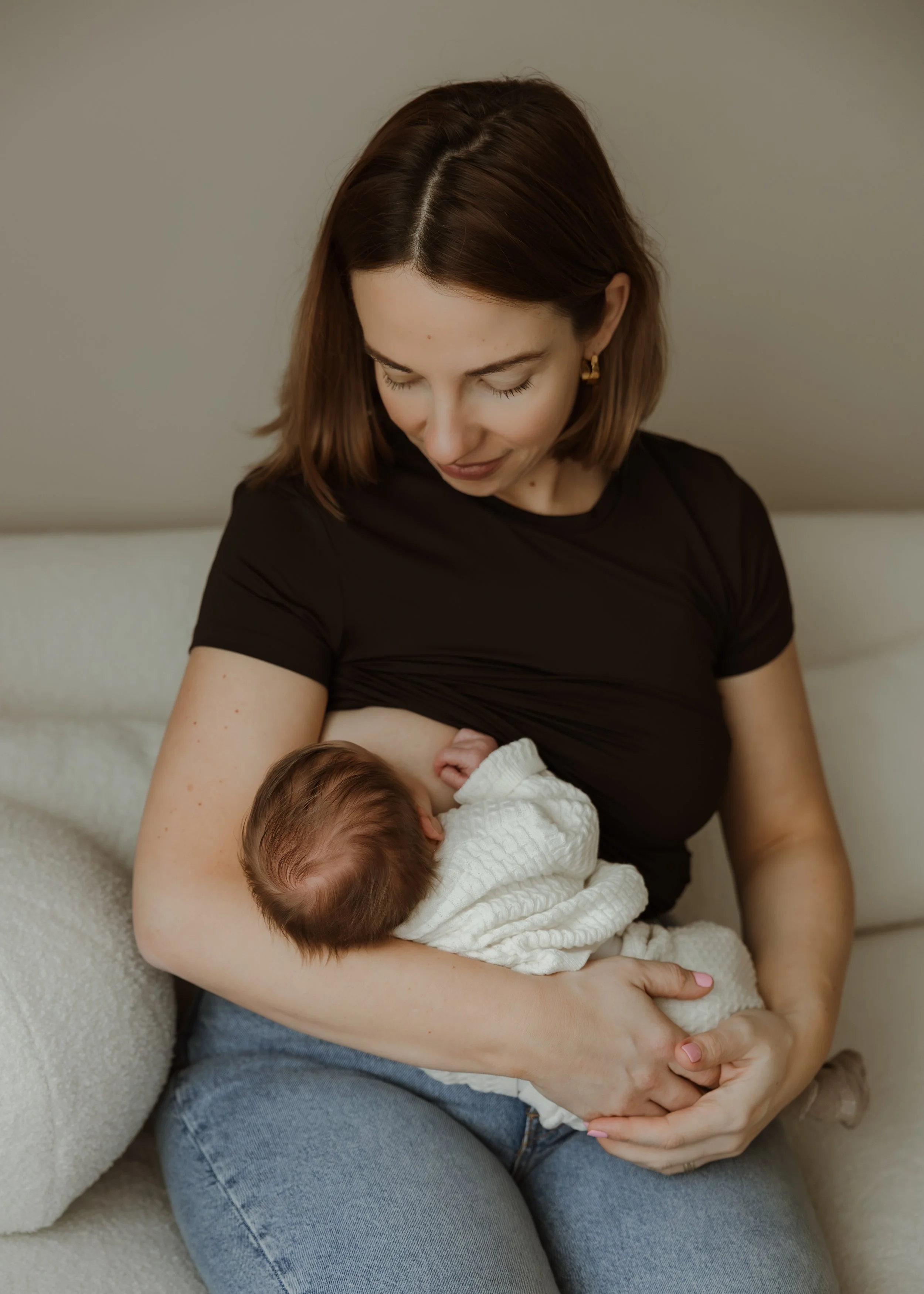 A woman with short brown hair wearing a black shirt and blue jeans is sitting on a white couch, gently holding a newborn baby wrapped in a white blanket, breastfeeding the baby.