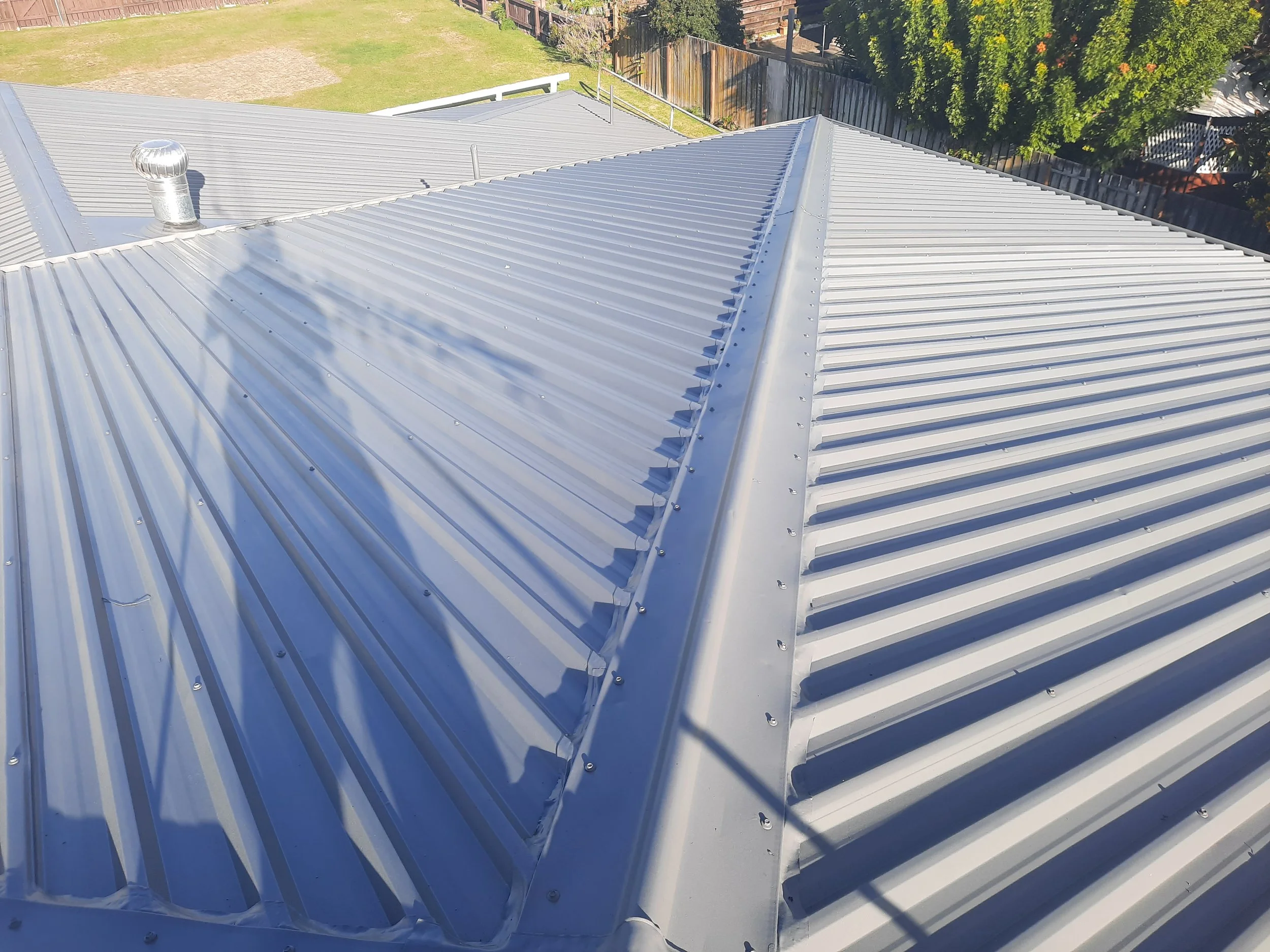 White corrugated metal roof with a roof vent, viewed above a fenced backyard and lawn