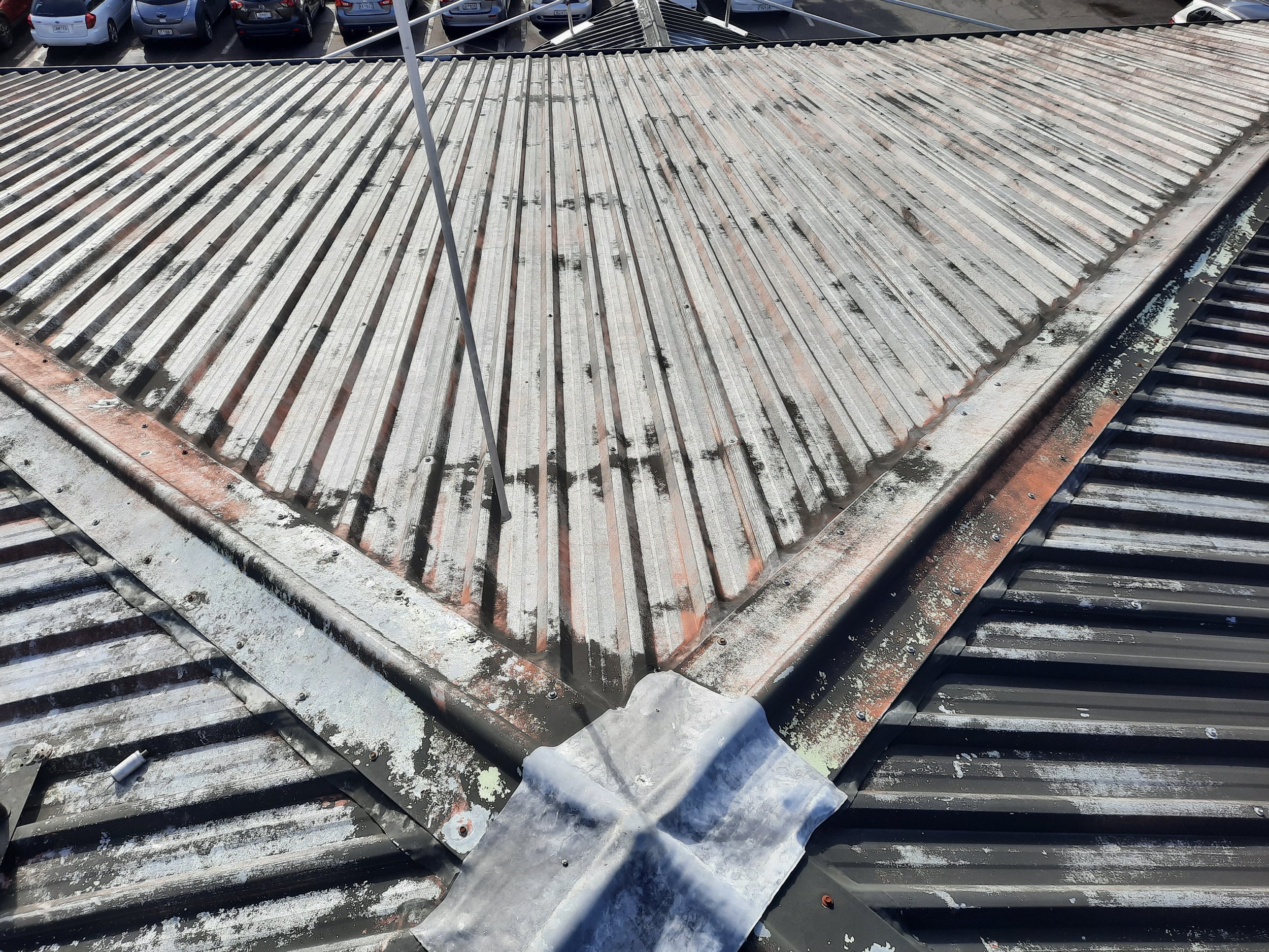 Weathered corrugated metal rooftop with a central roof vent, rusted flashing, and parked cars visible below