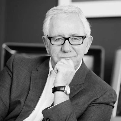 An older man with gray hair, glasses, and a suit, sitting in an office chair with a thoughtful expression.