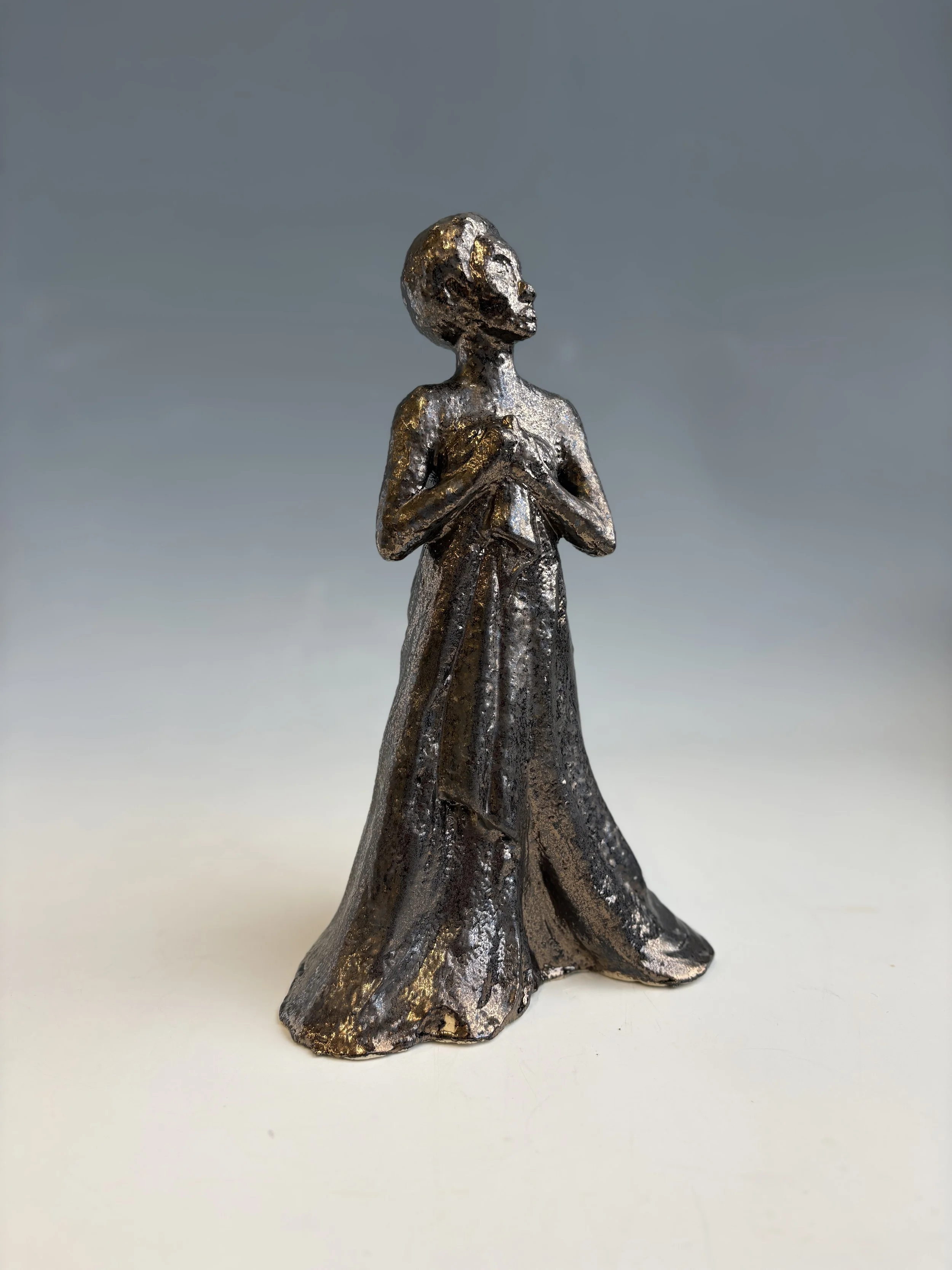 Metallic figurine of a woman with short hair, holding her chest with both hands, standing on a flat surface against a cloudy sky background.