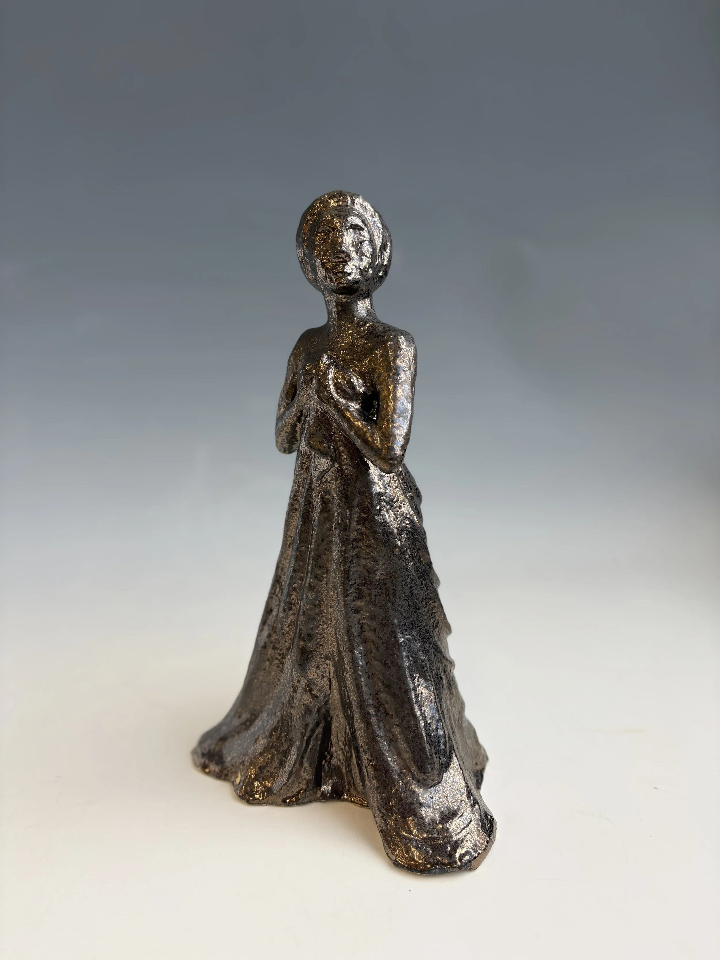 Metallic figurine of a woman in a flowing dress, with one hand on her chest and the other arm resting at her side, standing against a gray gradient background.
