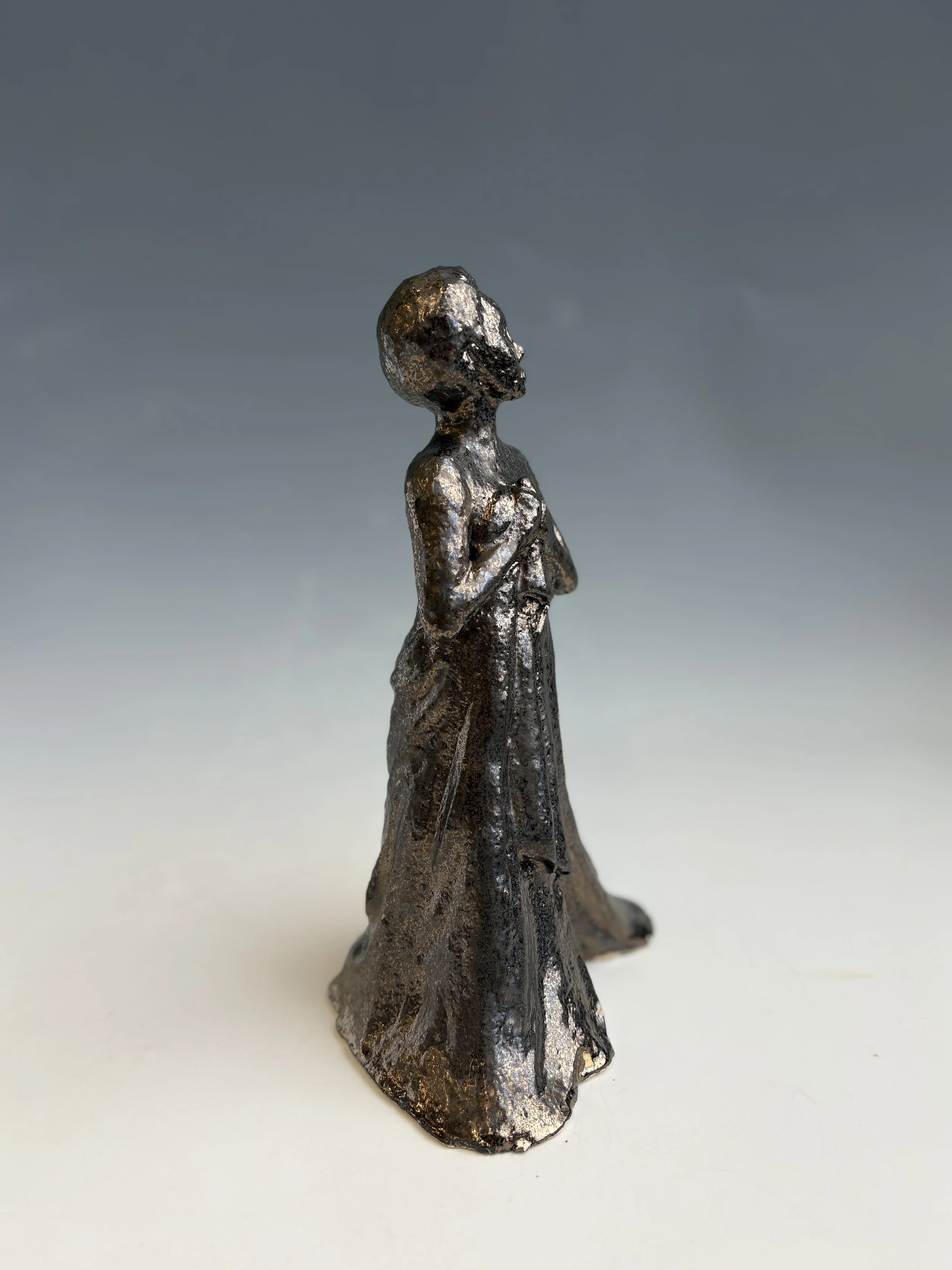 Metallic sculpture of a woman with arms crossed, standing on a white surface with a plain gray background.