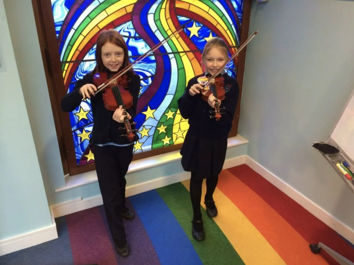 Two pupils playing violin in front of a stained-glass window