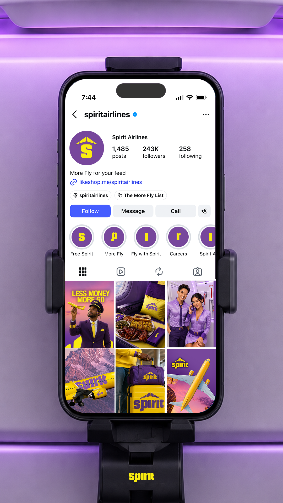 A smartphone mounted on a stand displaying the Instagram profile of Spirit Airlines. The profile has a purple and yellow logo, with several highlighted story icons and recent posts related to the airline's branding and promotions. The background has purple lighting.