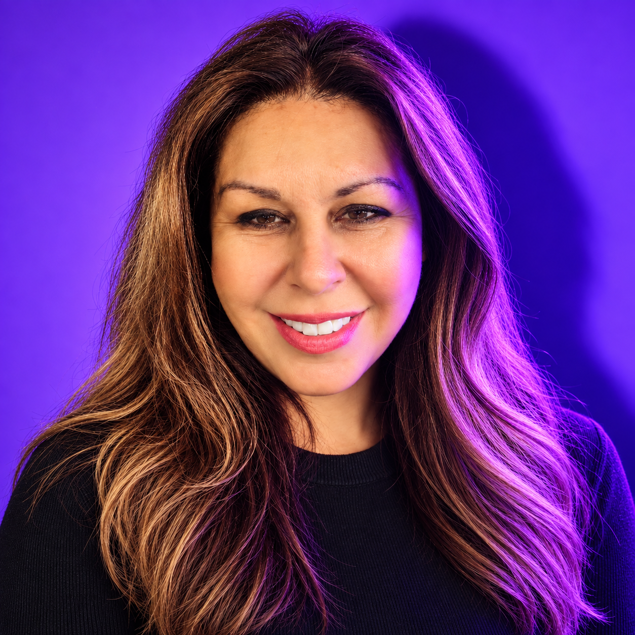 A woman with long, wavy brown hair smiling, wearing a black top, with purple lighting in the background.