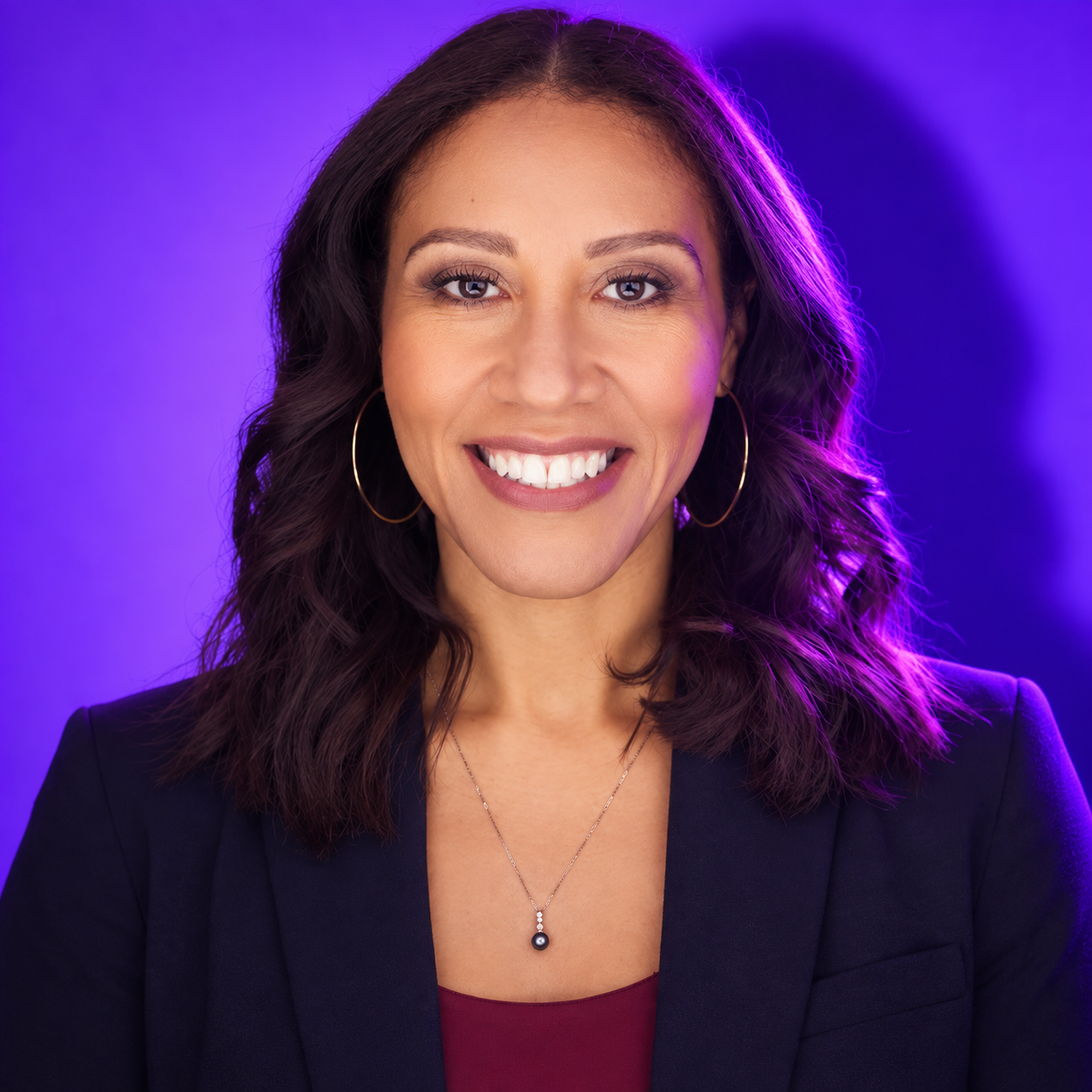 A woman smiling with shoulder-length wavy dark hair, wearing hoop earrings, a dark blazer, and a necklace, against a purple background.